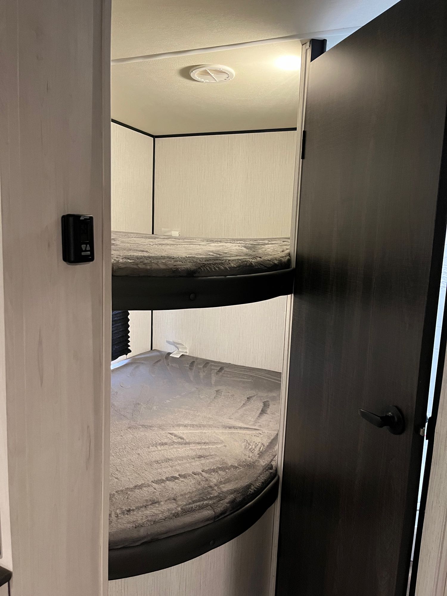 An RV rental features two cozy bunk beds with grey bedding. A thermostat is on the left wall, and a dark door is ajar on the right.. Heartland RVs Trail Runner 2022