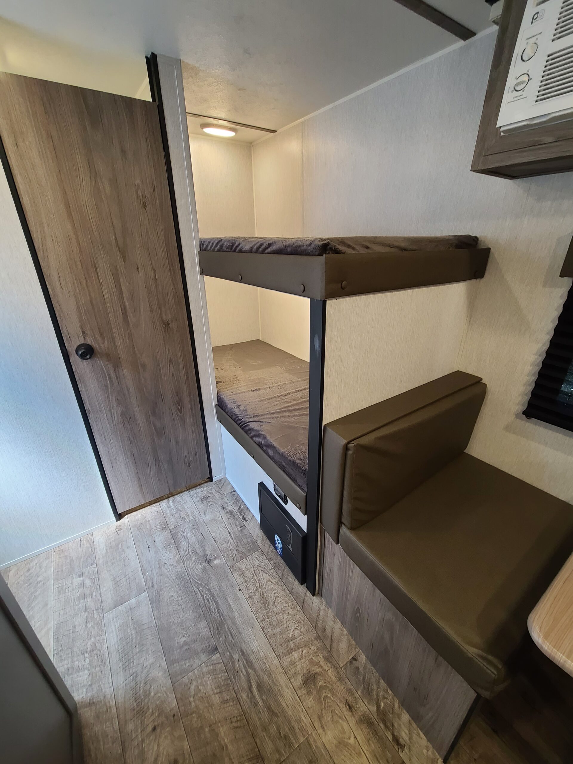 Inside an RV rental: two bunk beds with dark mattresses, a brown dinette bench, wood-look flooring, and a wooden door.. Sunset Park RV Sun Lite LTD 2024