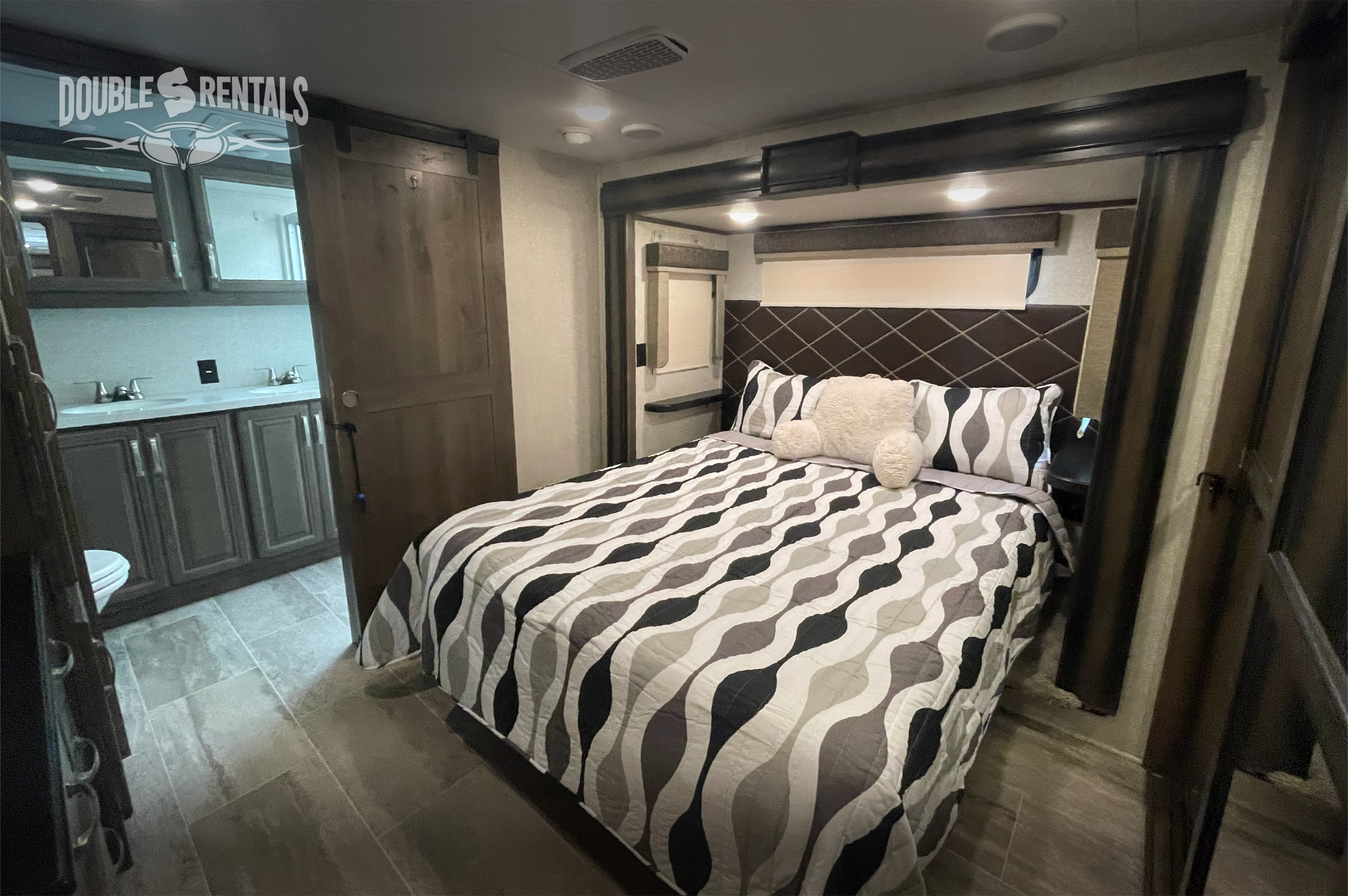 Step inside this cozy RV rental's master bedroom, complete with a modern bed and an en-suite bathroom visible through a barn door.. Keystone Montana 2019