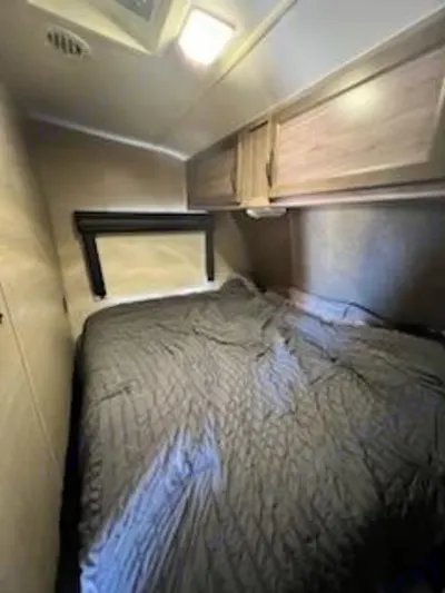 RV rental bedroom: dark bedding on a bed, light wood overhead cabinets, and a headboard shelf with a window.. Pacific Coachworks Blaze'N 2017