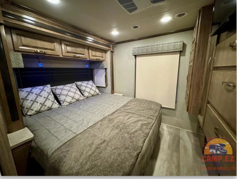 Modern RV rental bedroom with a large bed, dark headboard, overhead cabinets, blue accent lights, and light wood flooring.. Jayco Seneca 2020
