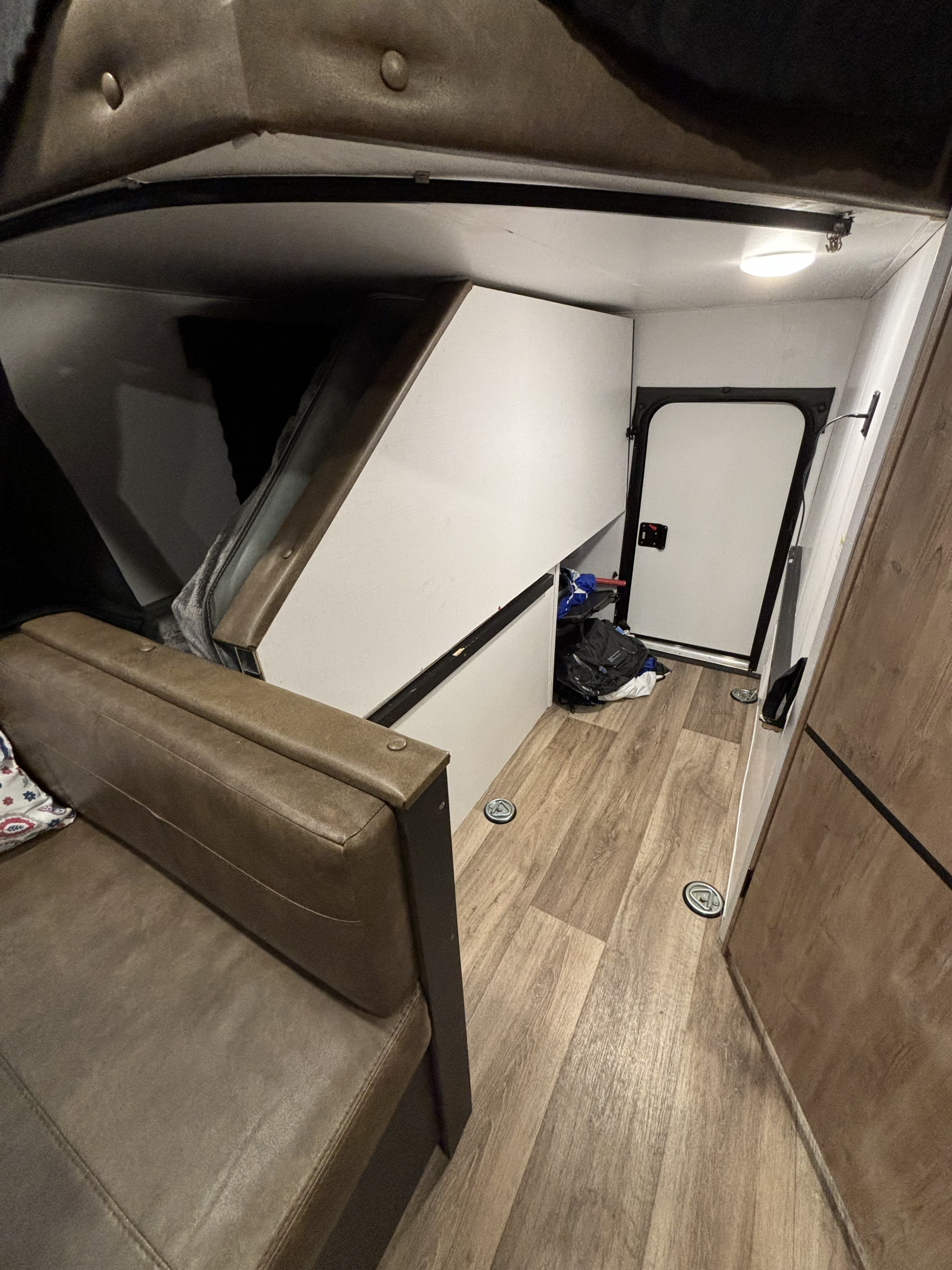 Interior view of an RV rental showing a brown sofa, a raised bunk bed area, wood flooring, and a side door.. Keystone RV Outback Ultra-Lite 2021
