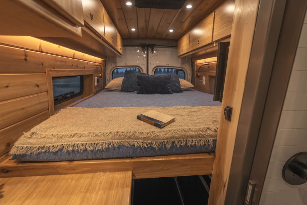RV rental interior: A bed with blue bedding and a book, set within a cozy wooden camper van with custom cabinets and warm lighting.. Ford Transit 2023