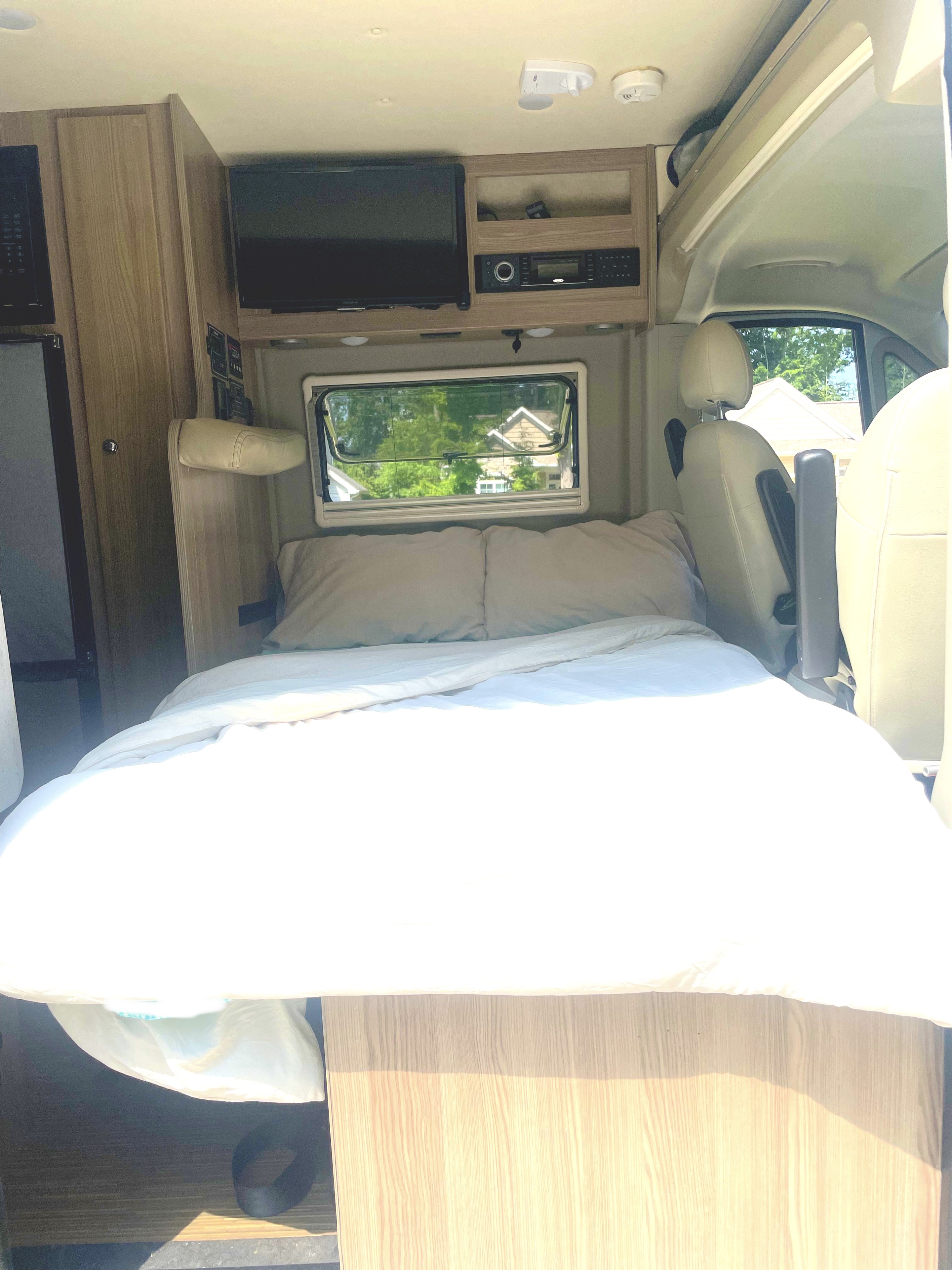 Inside a bright RV rental, a comfy bed is made with white bedding, beneath a window, TV, and wooden cabinets.. Winnebago Travato 2019