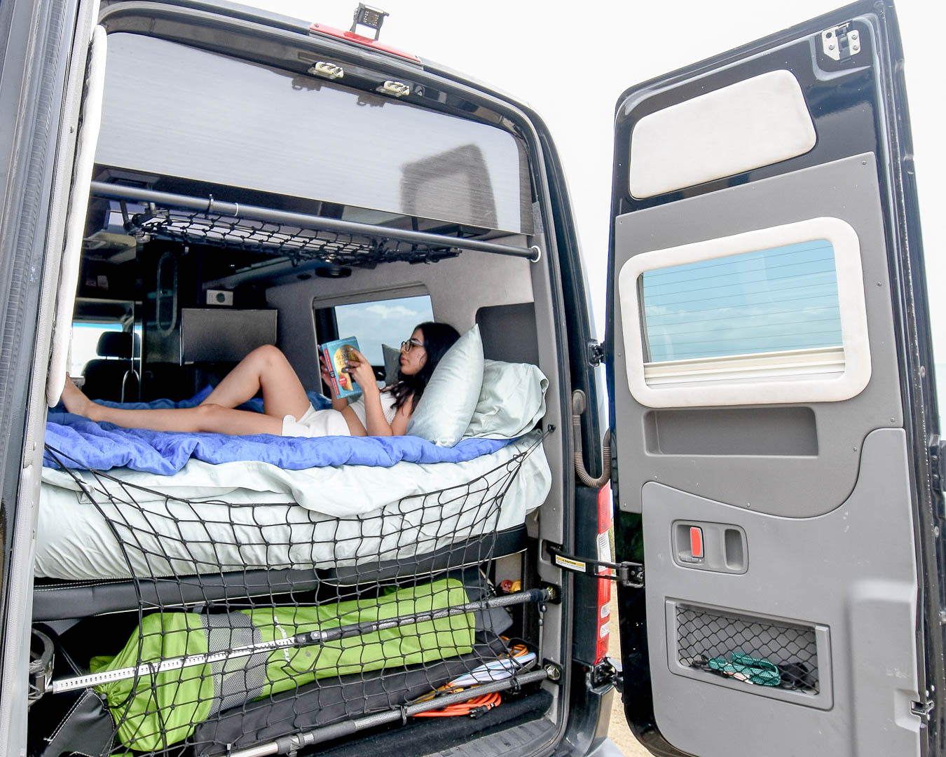 Enjoy an RV rental! A woman relaxes reading in a spacious camper van with open rear doors, revealing cozy bed & storage.. Airstream Interstate 2015