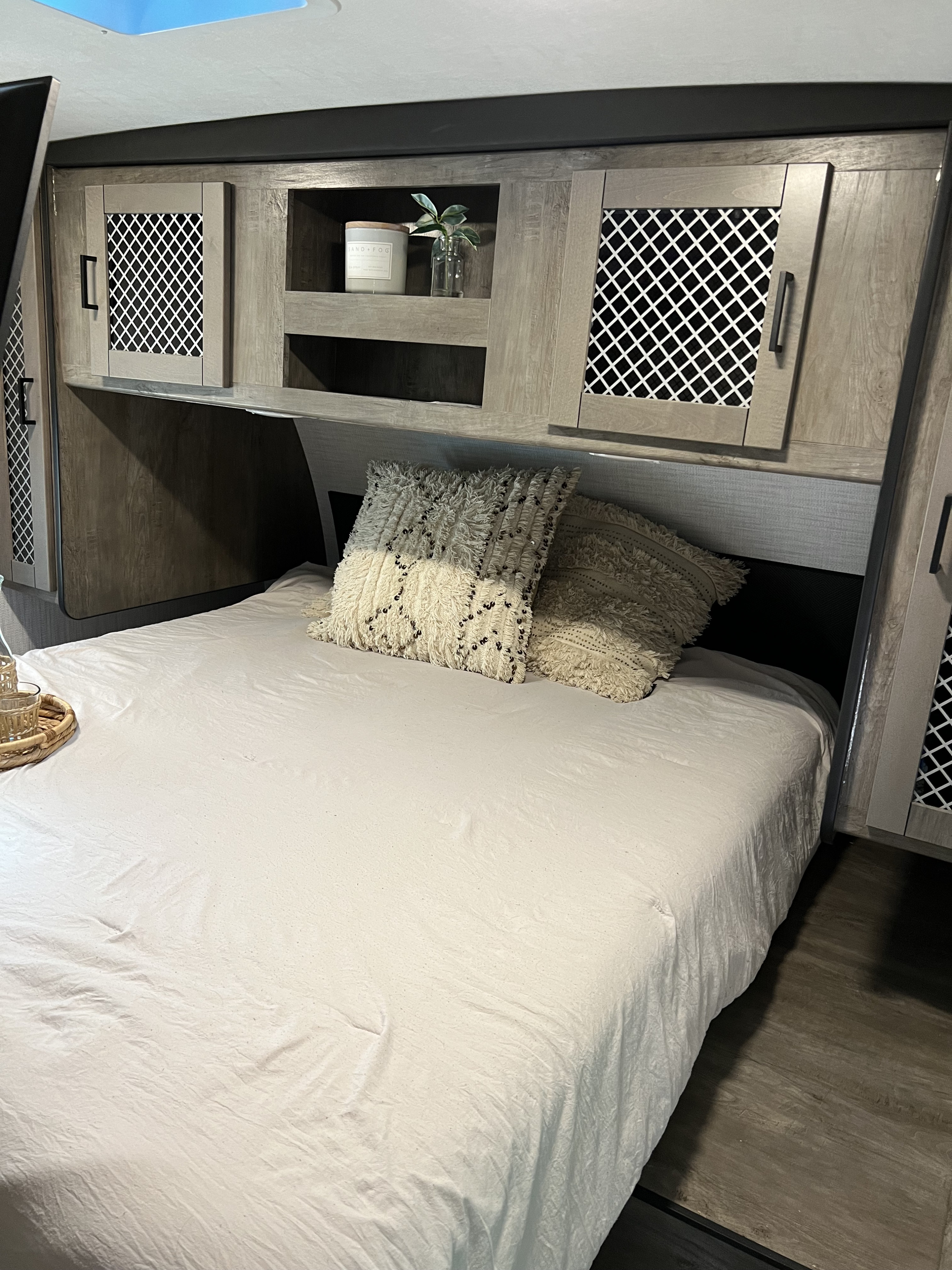 This RV rental bedroom features a cozy bed with white linens and patterned pillows, overhead cabinets, and built-in shelving.. Heartland RVs North Trail 2021