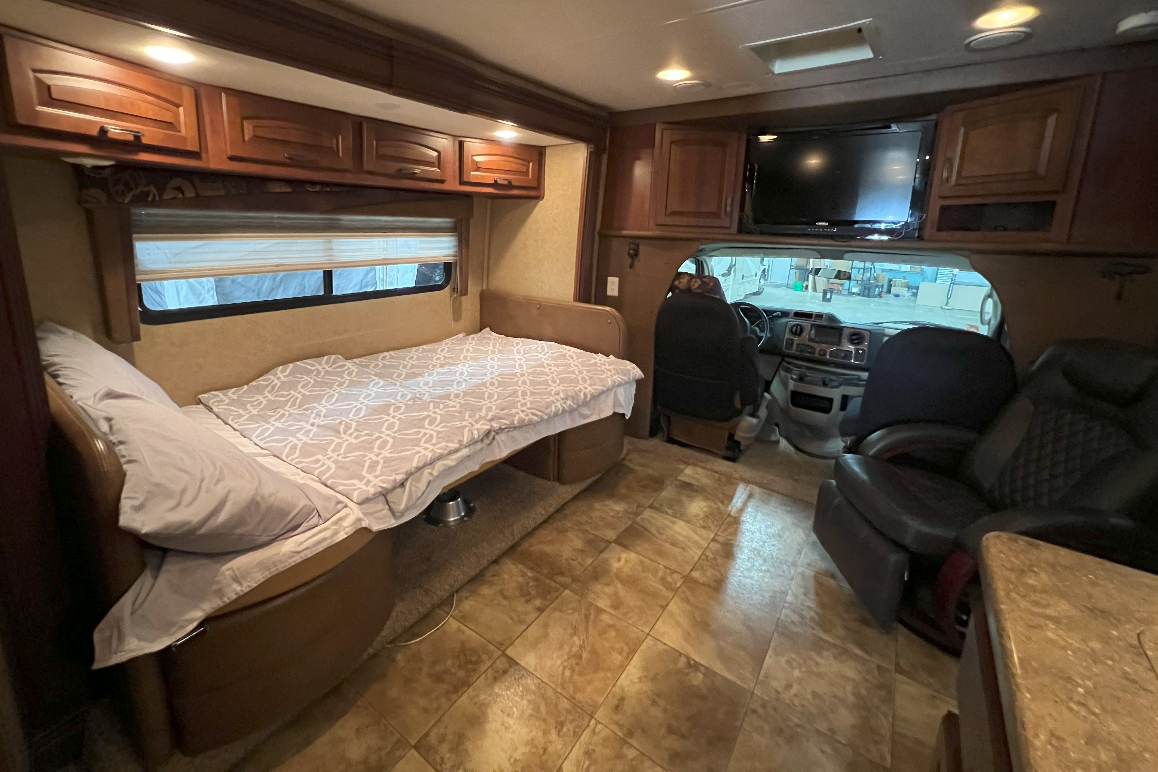 The interior of an RV rental: a comfortable bed on the left, driver's cockpit visible ahead, TV, and overhead cabinets.. Jayco Melbourne 2013