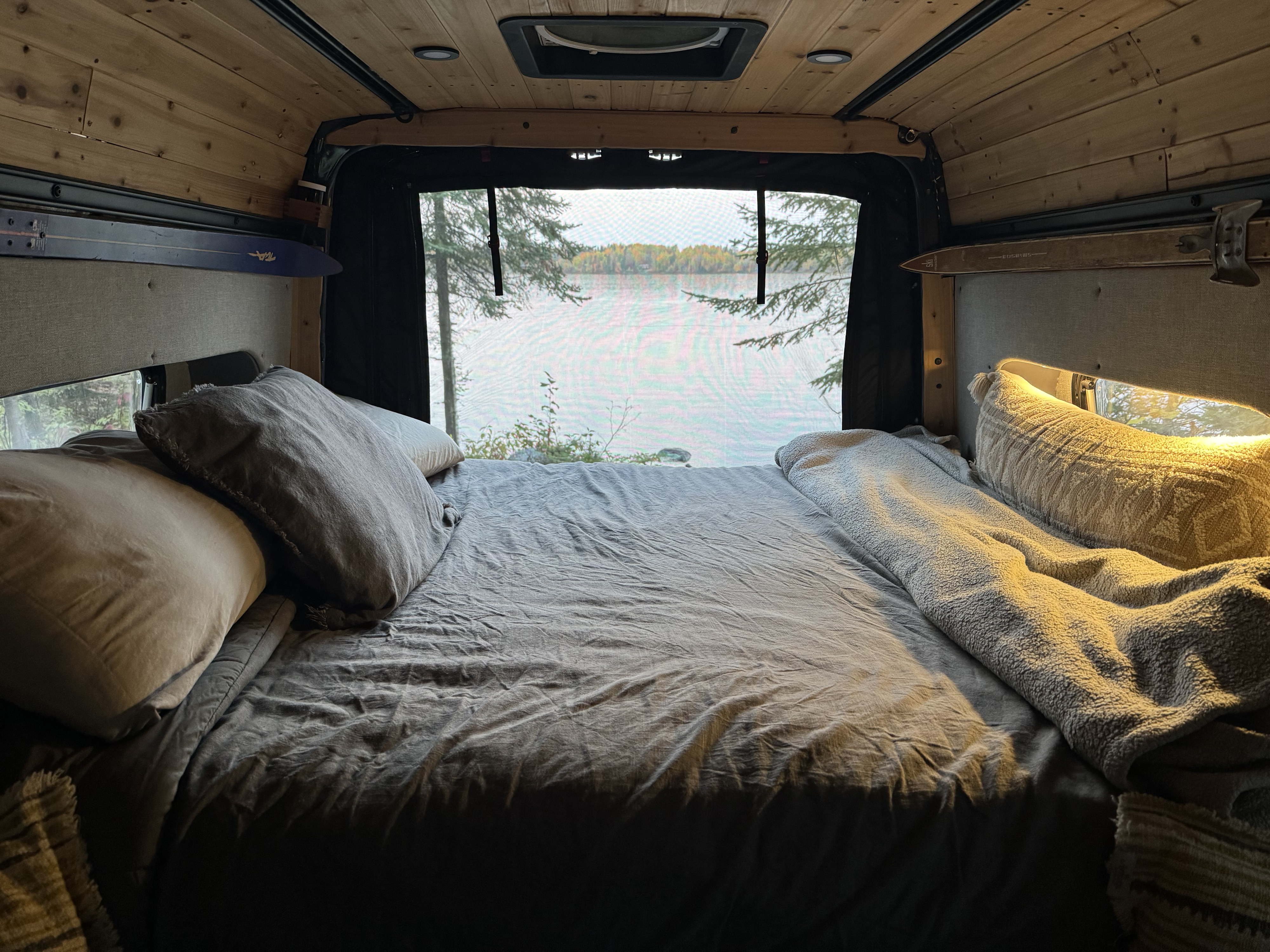 Cozy RV rental bed with a serene lake view and autumn trees. Perfect for peaceful van life getaways.. Mercedes Sprinter Van 4x4 2020