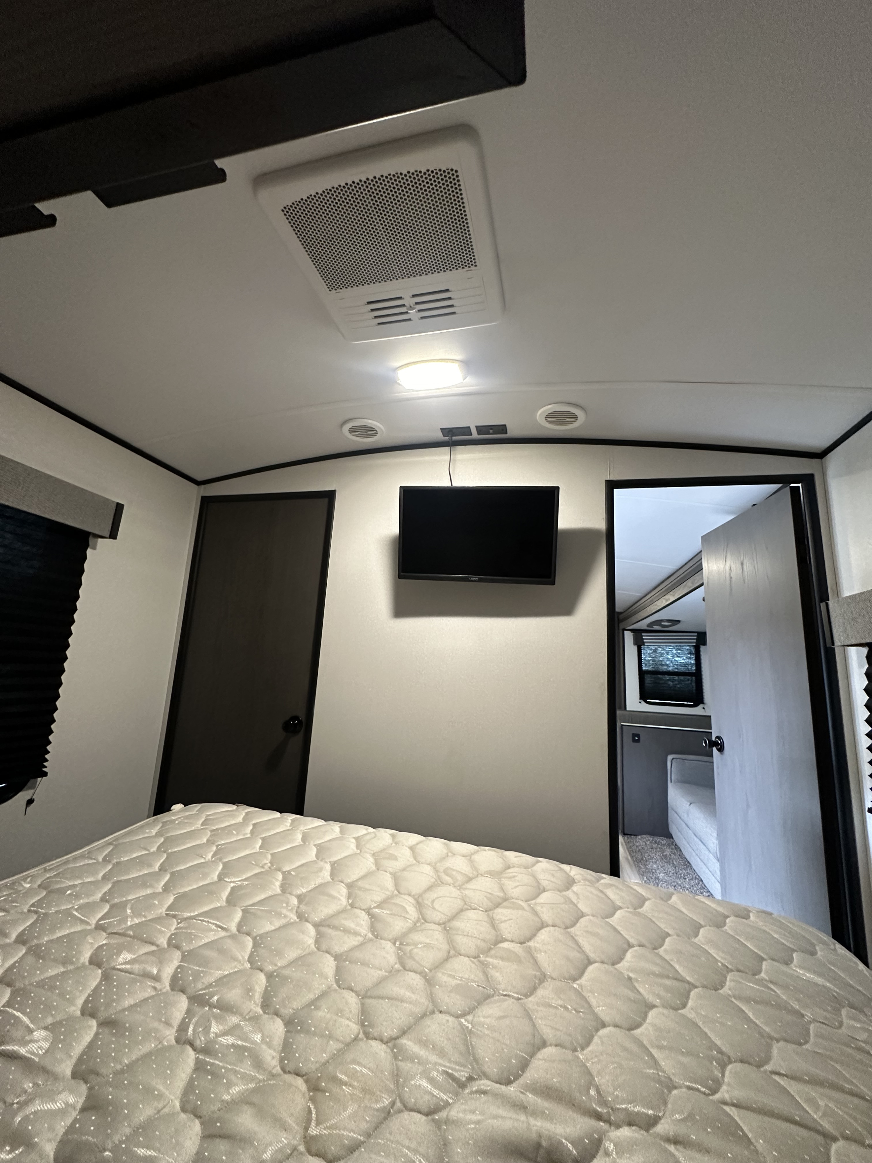 Inside an RV rental: a cozy bedroom with a quilted mattress, wall-mounted TV, dark doors, and overhead AC unit.. Heartland RVs MPG 2800QB 2021
