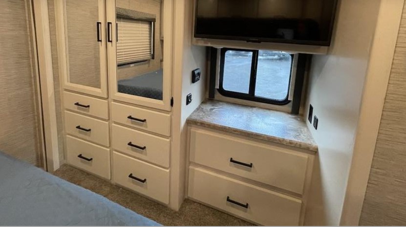 Explore this efficient RV rental bedroom setup: mirrored storage cabinets, ample drawers, TV, and a window view.. Thor A.C.E. 2023
