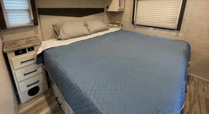 RV rental bedroom featuring a comfy bed with blue quilted cover, built-in nightstands, and windows with blinds.. Thor A.C.E. 2023