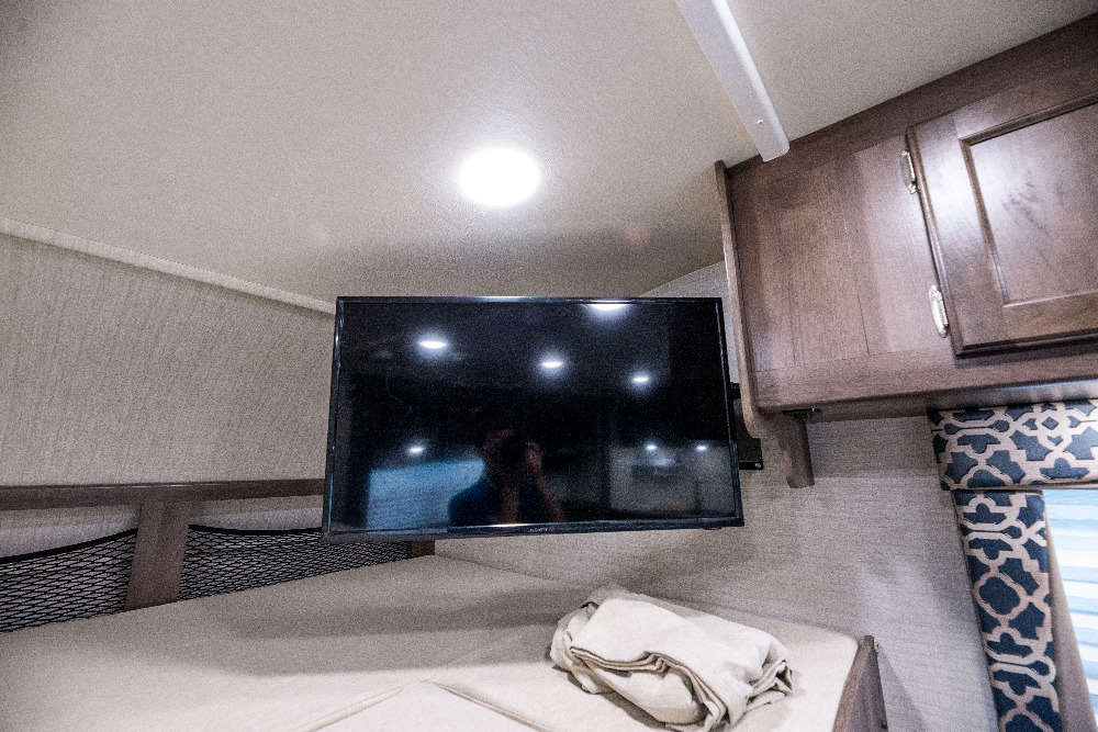 RV rental sleeping area with mounted TV above the bed, featuring overhead cabinets and patterned window details.. Jayco Redhawk 2018