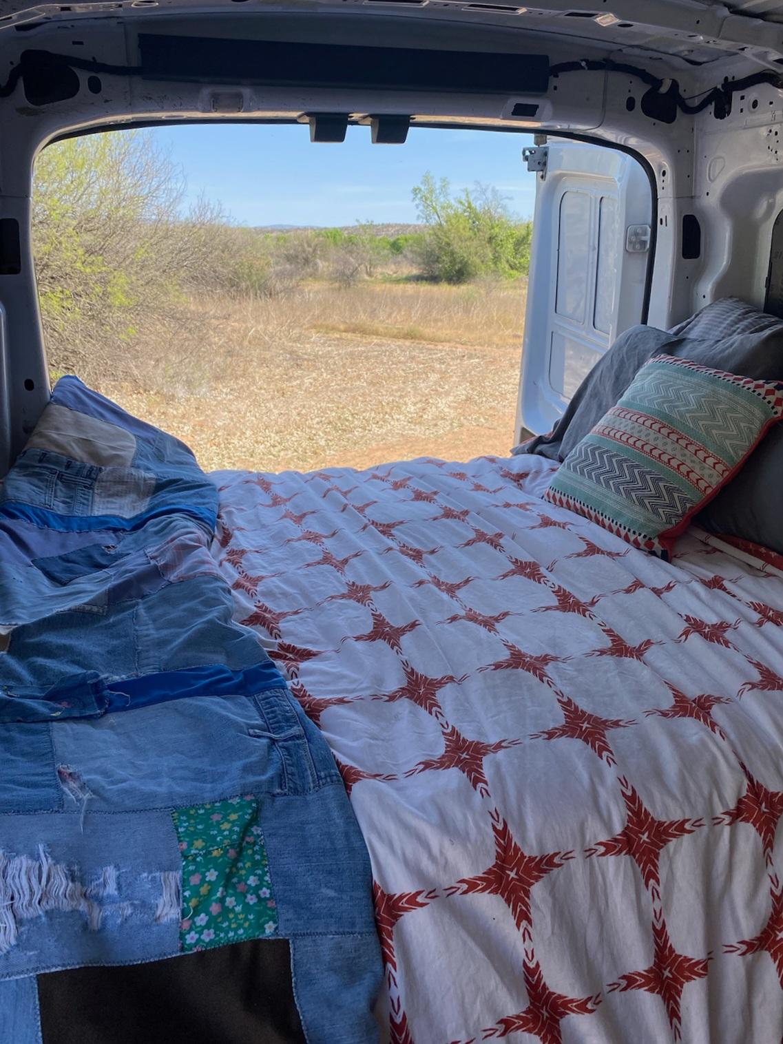 Cozy bed in an RV rental, with open back doors revealing a dry, natural landscape under a clear sky.. FORD Ford Transit 2015