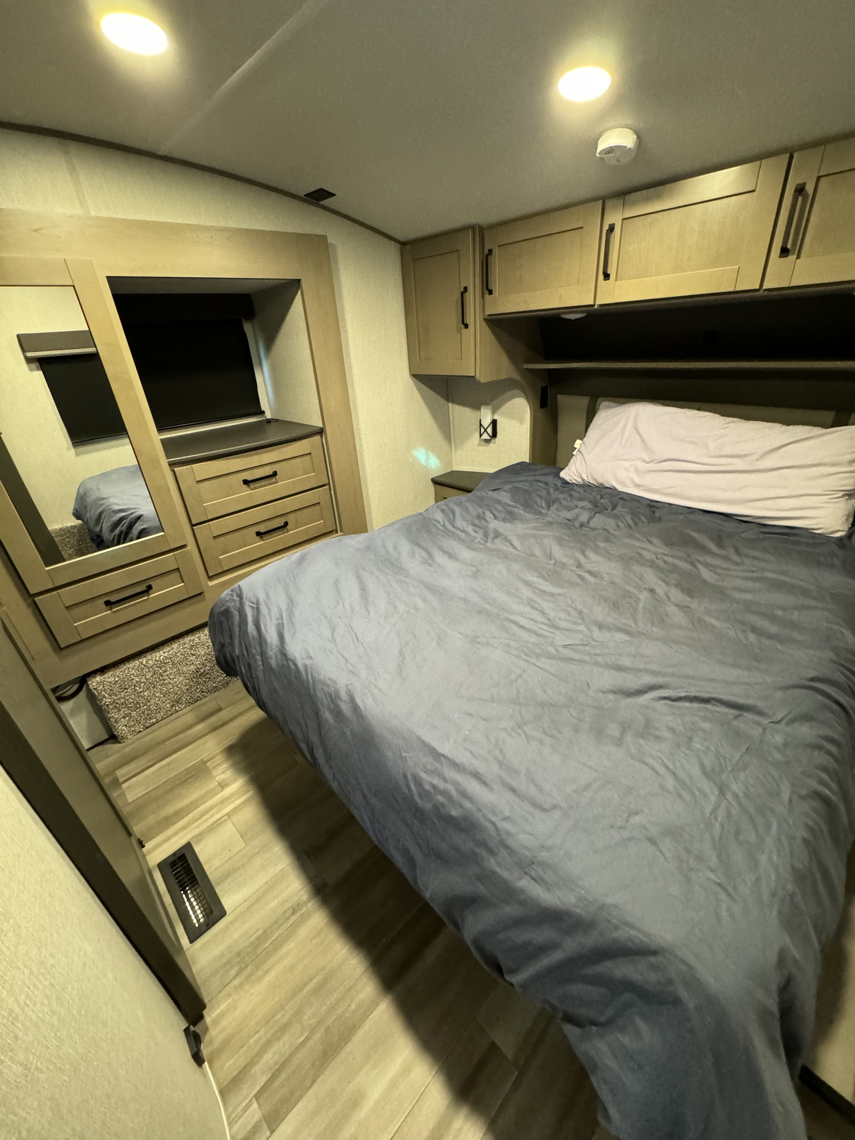 This cozy RV rental bedroom features a comfy bed with blue bedding, ample storage, and stylish wood-look flooring.. Grand Design Reflection 2024