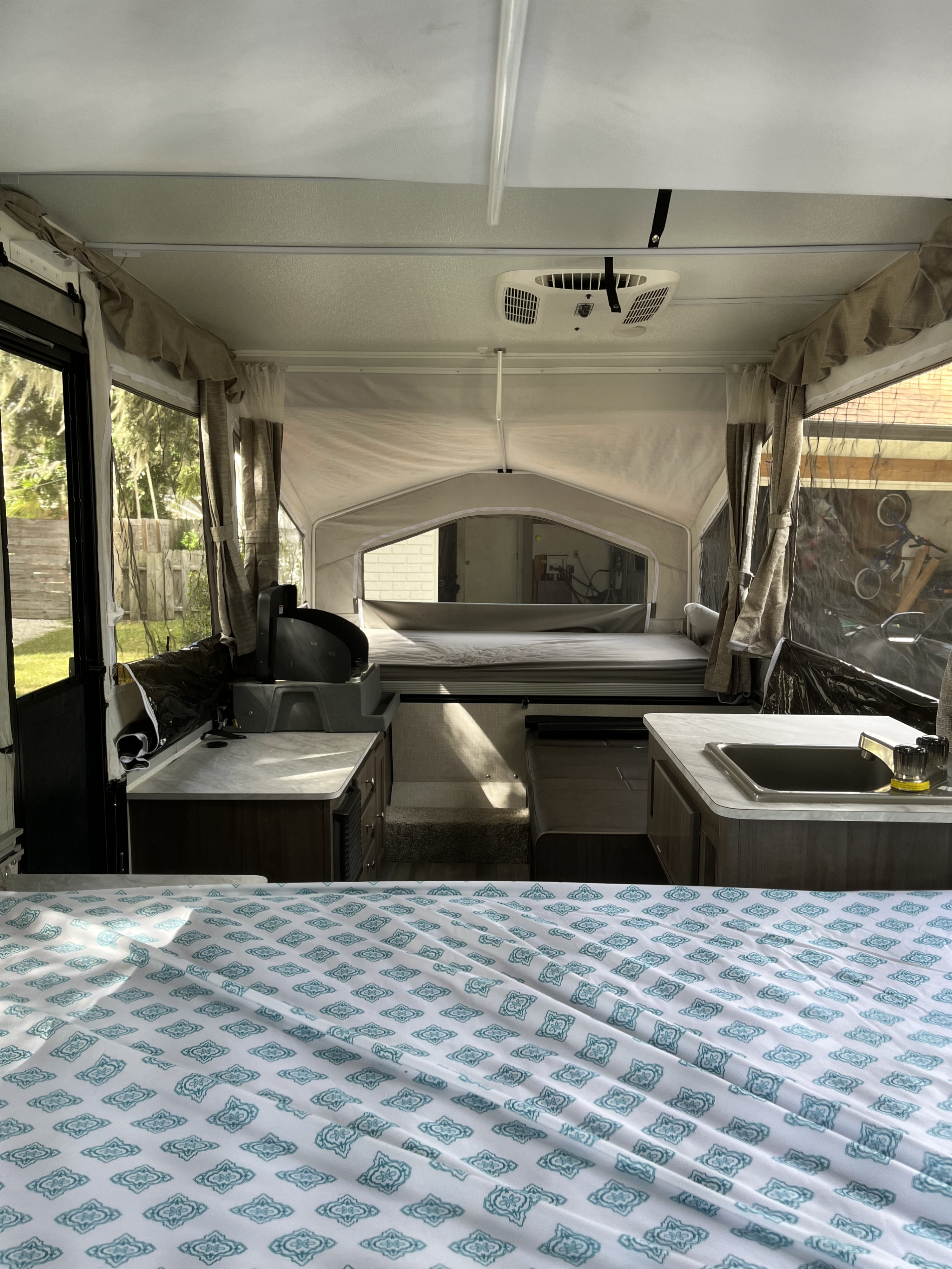 Inside a pop-up RV rental, foreground bed with patterned sheet, compact kitchen with sink/stove, and back bed visible.. Forest River Flagstaff LTD 2023
