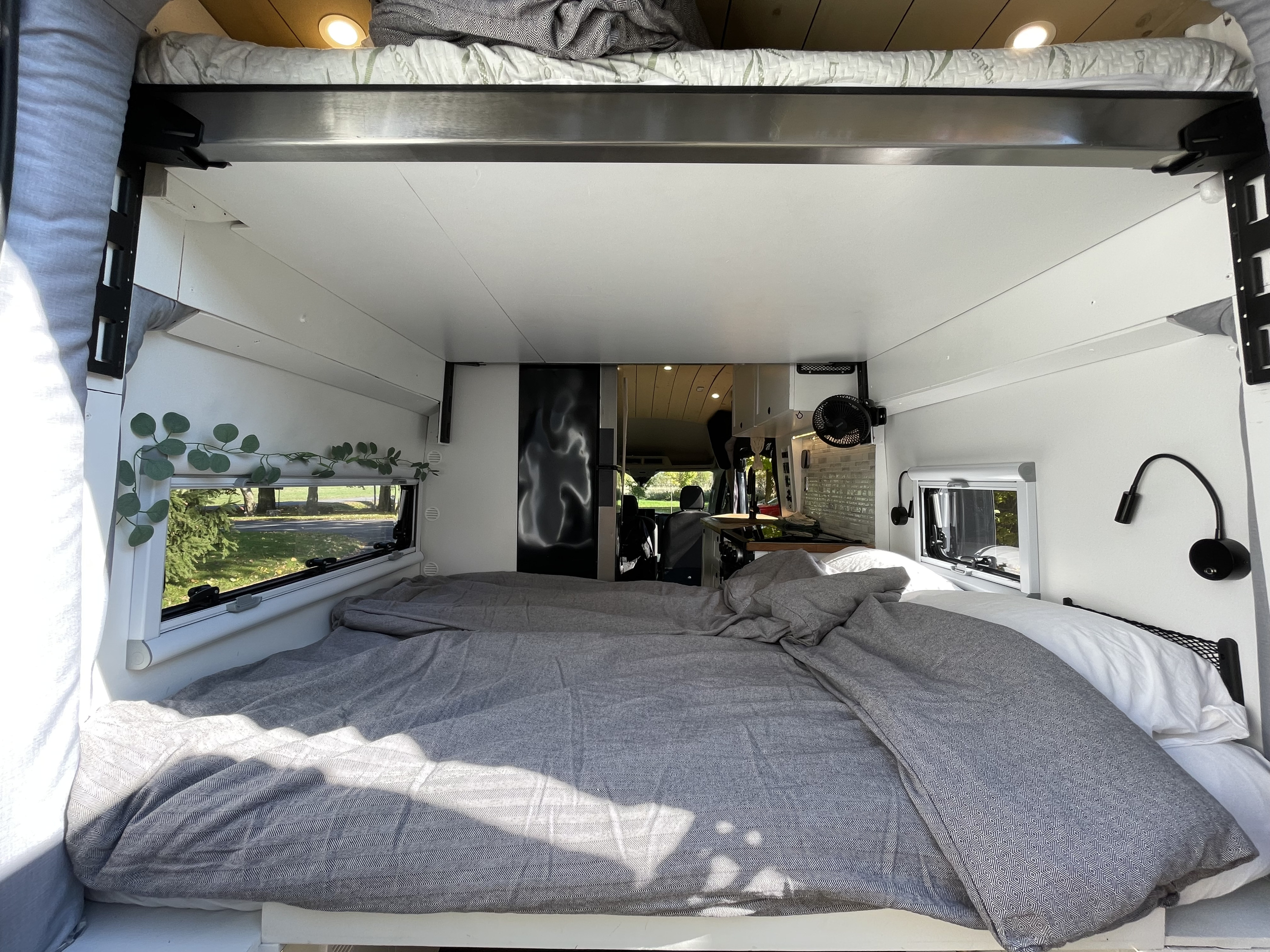 Experience comfort in this RV rental! Features a large, made bed with grey bedding, side windows, and a view into the kitchen.. Ford Transit 2023