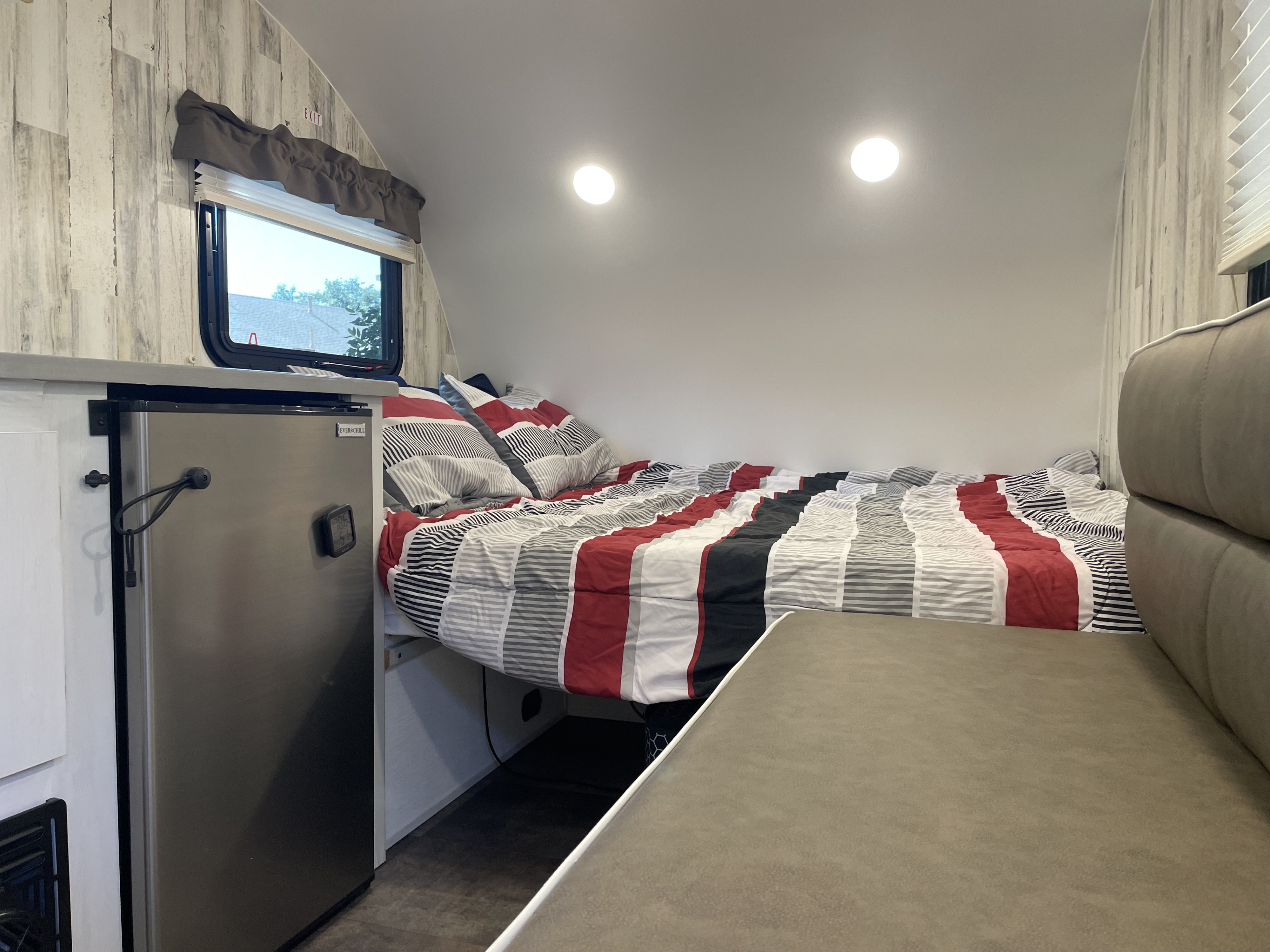 An RV rental interior features a bed with striped bedding, a stainless steel mini-fridge, and an "EXIT" sign near the window.. Travel Lite Rove Lite 2023