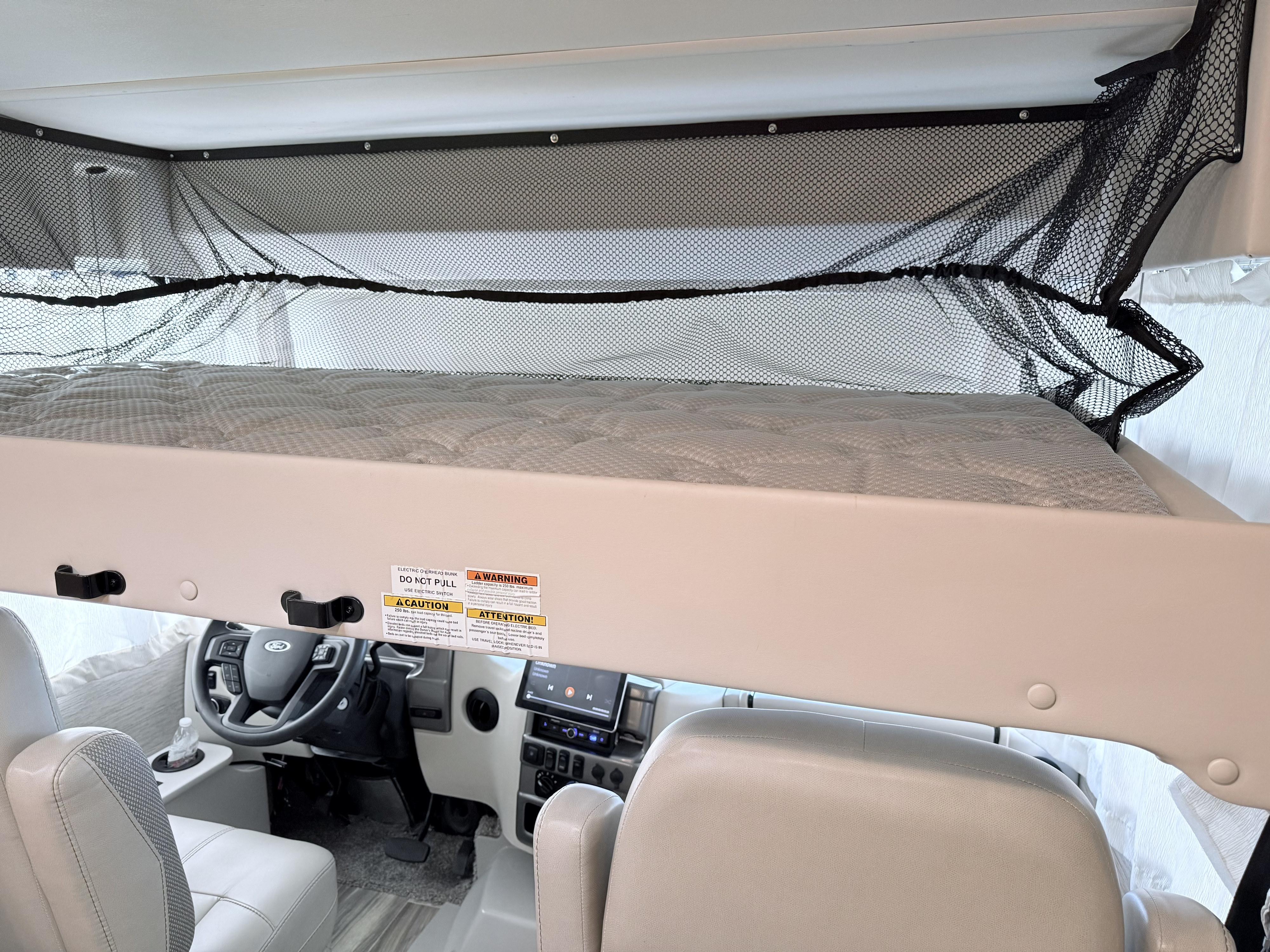 RV rental interior shows an overhead bunk bed with a safety net, positioned above the driver's and passenger's seats.. Thor Vegas 2023