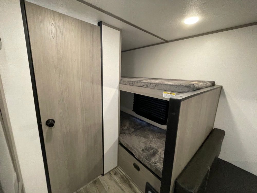 RV rental bunkhouse interior: Light wood door next to two stacked bunk beds with mattresses, a compact sleeping area.. Dutchmen Coleman Lantern 17B 2024