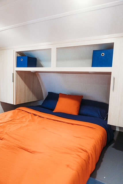 Modern RV rental bedroom with bright orange bedding, navy pillows, and ample overhead wood storage, including blue bins.. Forest River Tracer 2024