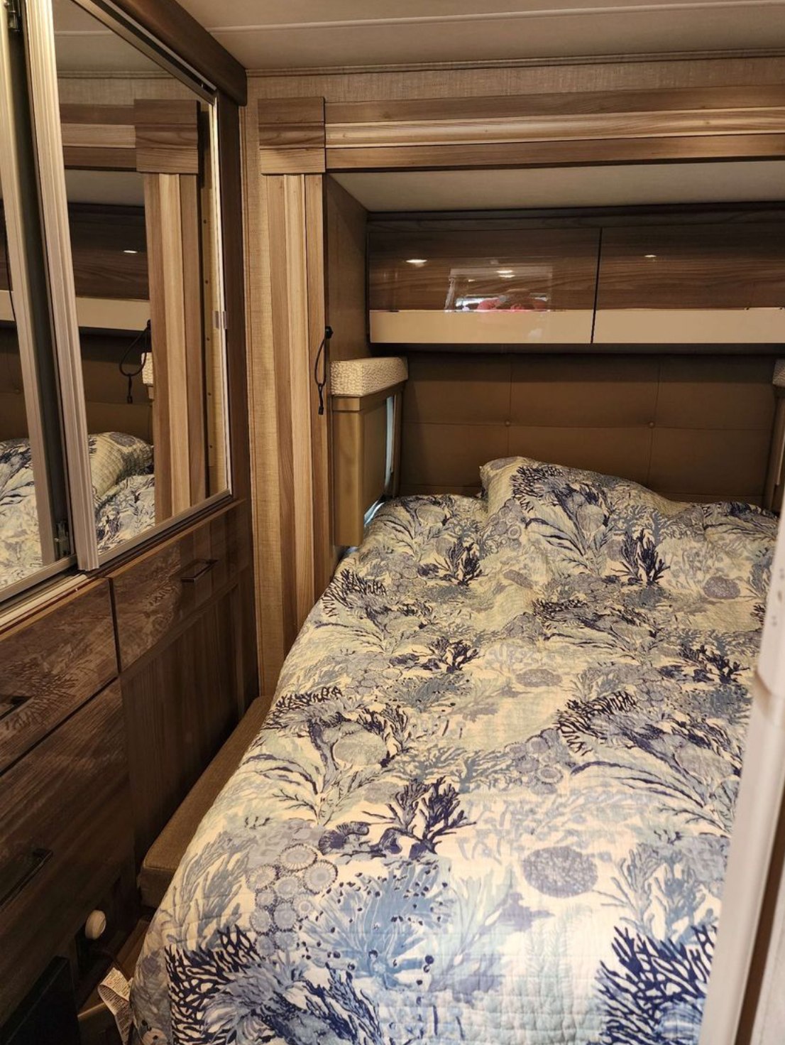 Cozy RV rental bedroom features a blue coral-patterned bedspread, mirrored wardrobe, and overhead storage, perfect for travel.. Thor Delano SV 2023