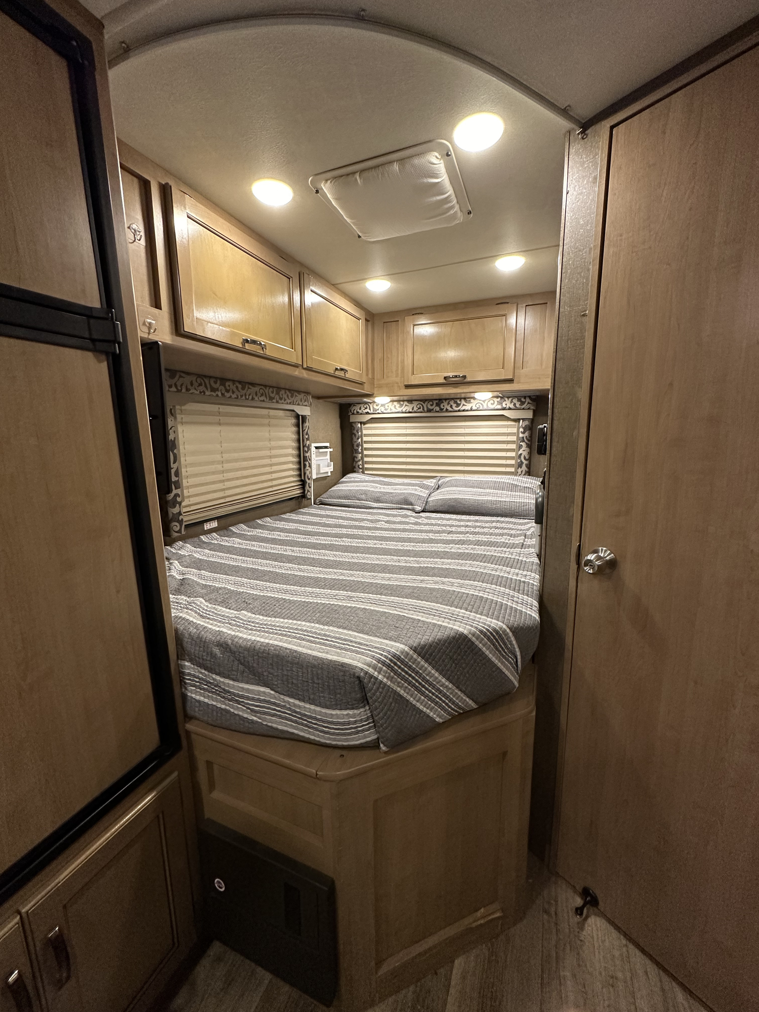 Comfortable RV rental bedroom featuring a striped queen bed, overhead cabinets, and bright interior lighting.. Thor Four Winds 2019