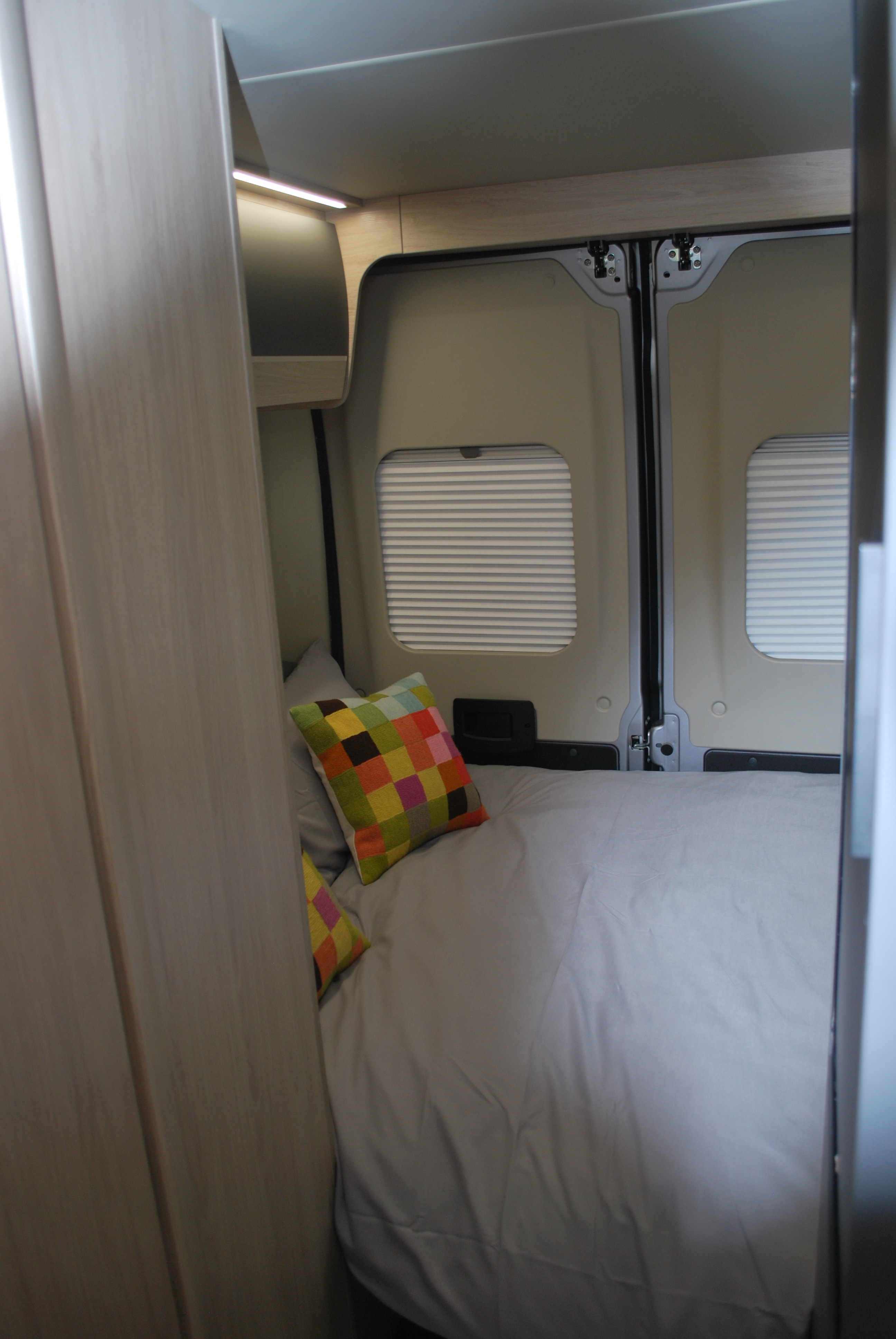 Cozy RV rental bedroom: bed with grey bedding, colorful pillows, rear windows with blinds, and overhead light.. Auto Trail Expedition 67 2024