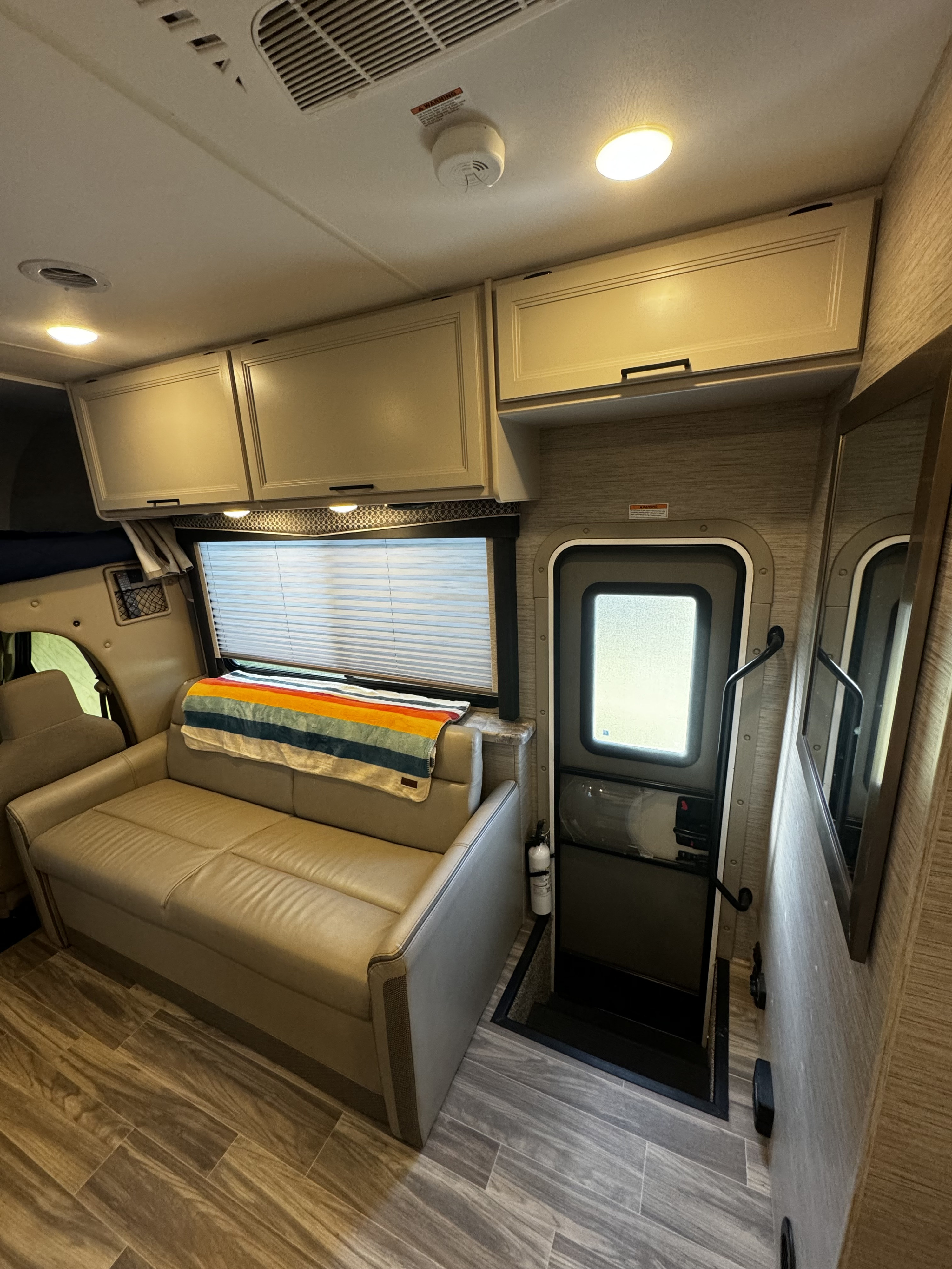 This RV rental interior features a cozy sofa, overhead cabinets, a door, and wood-look flooring. A fire extinguisher is visible near the entry.. Thor Motor Coach Four Winds 2022