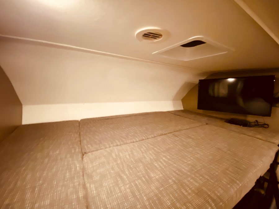 A cozy RV rental cab-over sleeping area with light-colored cushions, a wall-mounted TV, and a ceiling vent.. Winnebago 25 2019