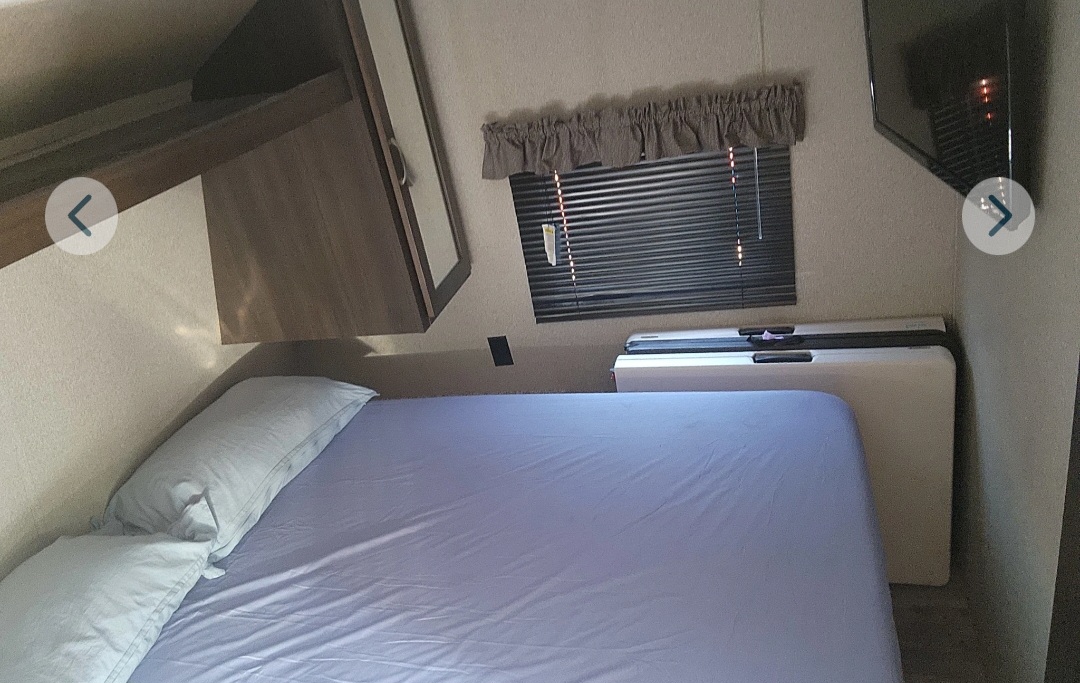 Inside an RV rental: a compact bedroom with a bed, wall-mounted TV, window, and overhead storage cabinets.. Keystone RV Springdale 2019