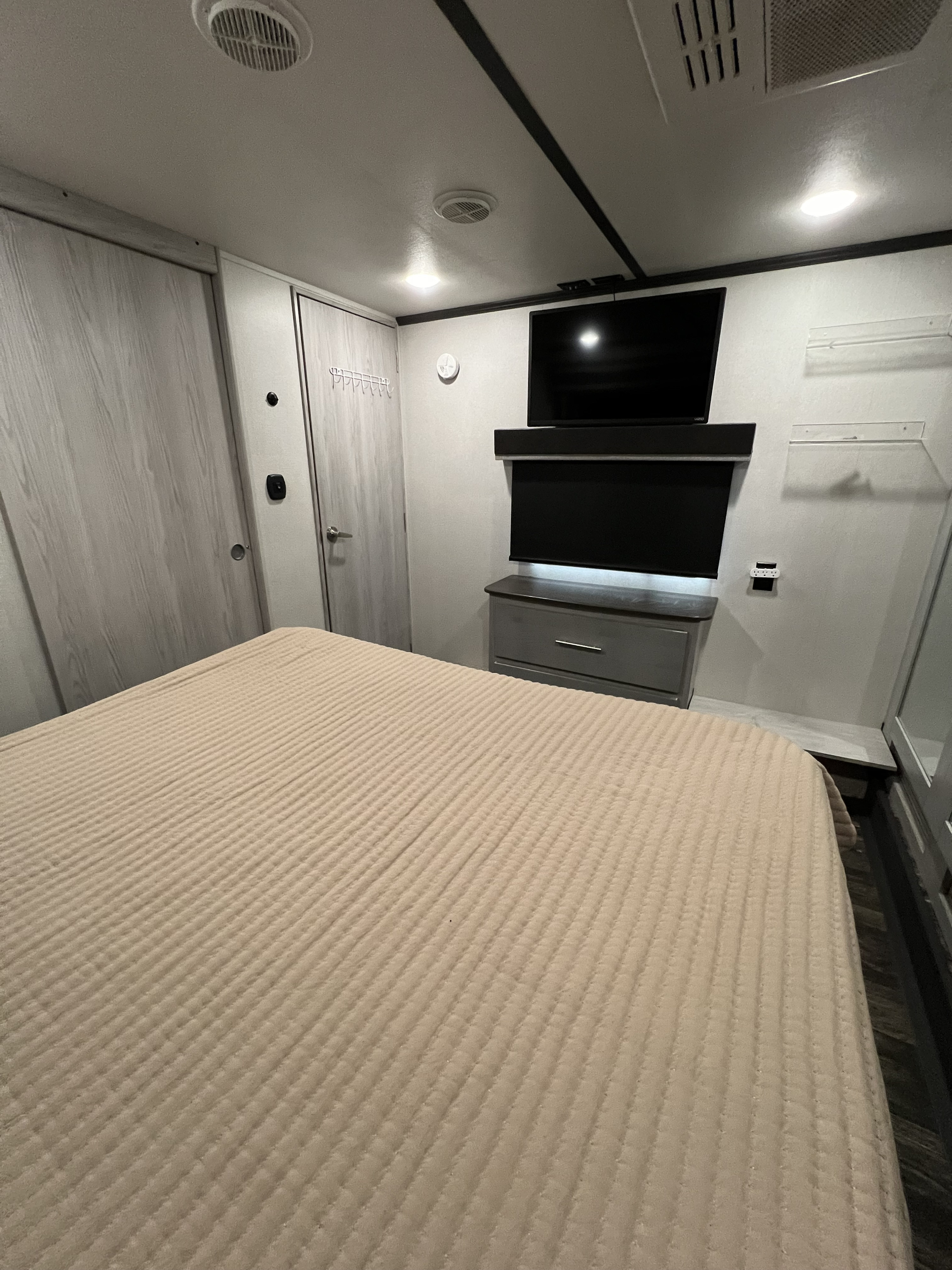 RV rental bedroom: Compact interior featuring a ribbed tan bed, gray dresser, wall-mounted TV, and light wood-grain closet doors.. Keystone RV Fuzion Toy Hauler 2021