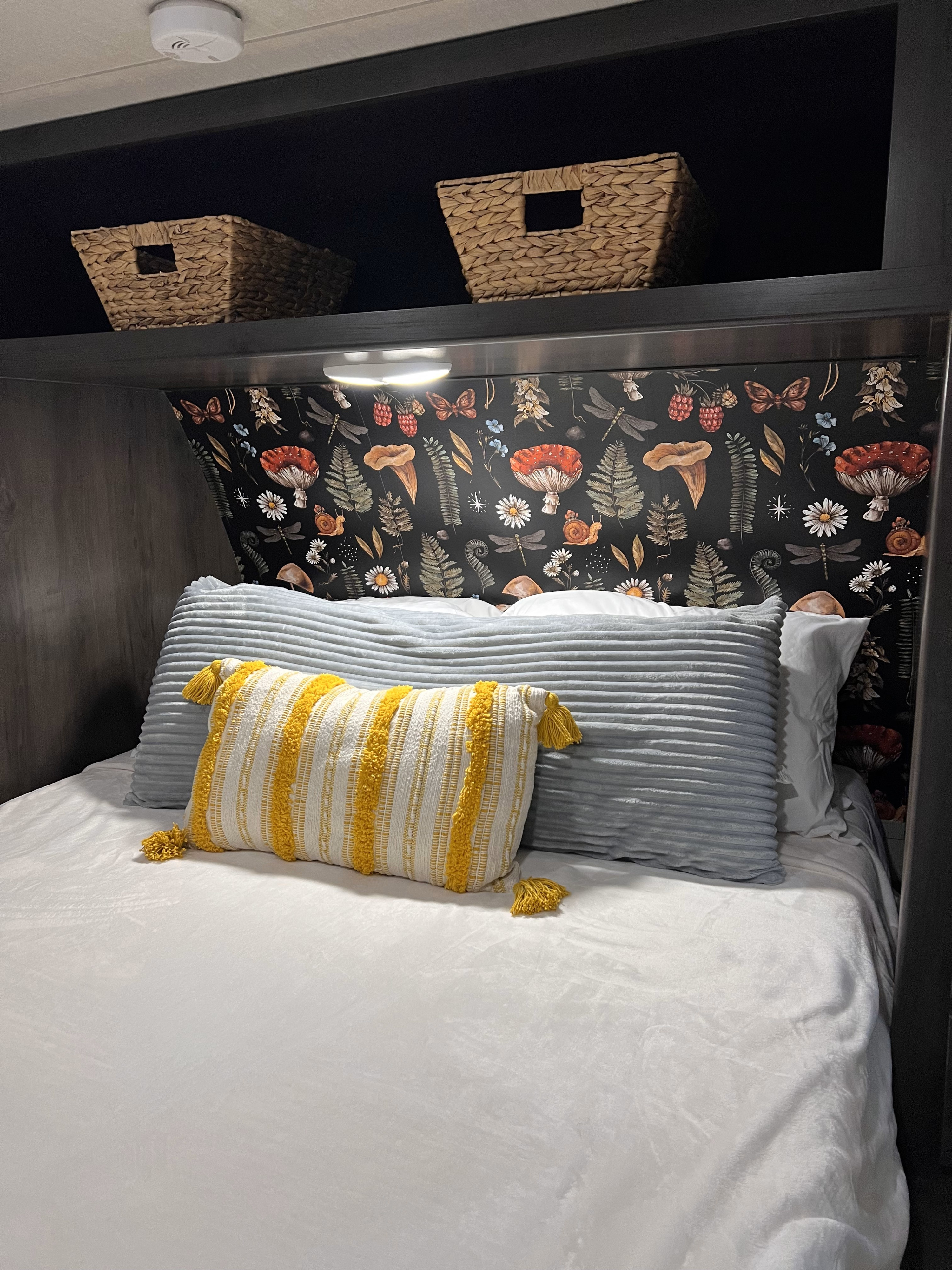 Cozy RV rental bedroom featuring a mushroom wallpaper headboard, comfy bed with pillows, and overhead storage.. Grand Design Transcend Xplor 2022