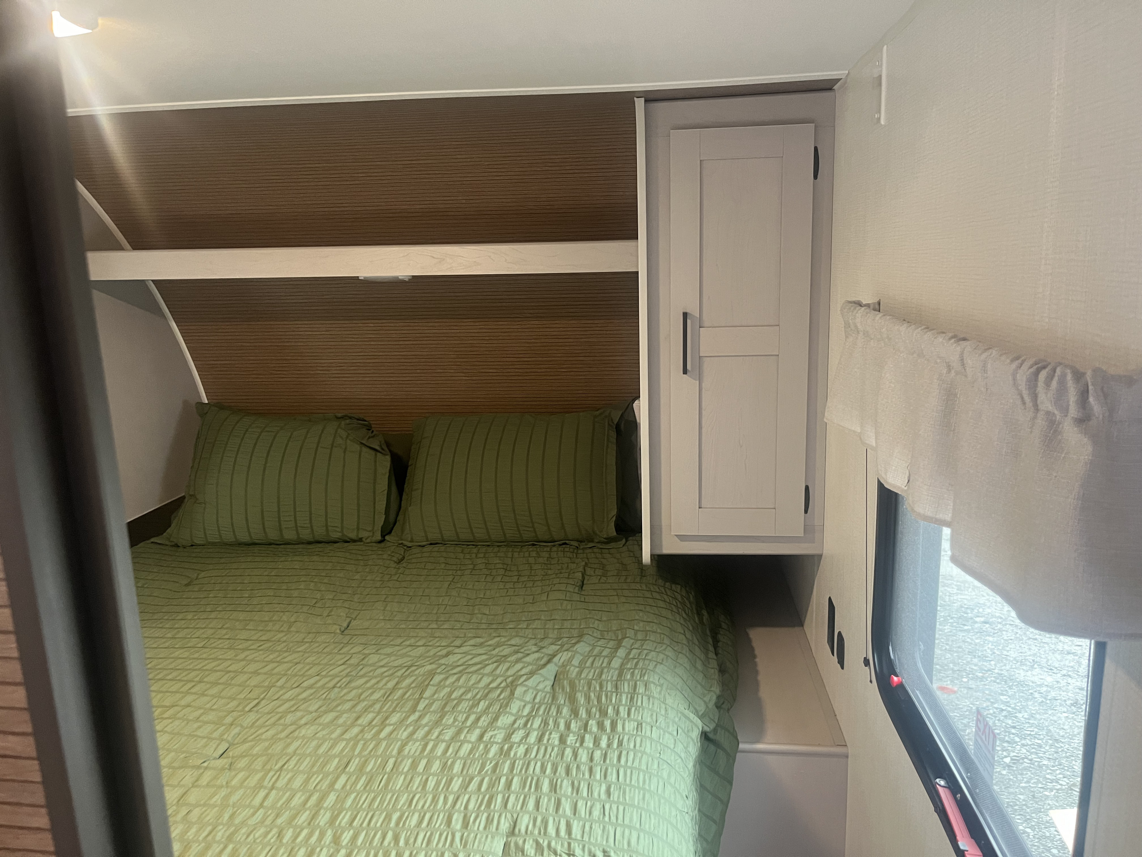 An RV rental bedroom, featuring a green-sheeted bed, light-colored overhead cabinet, and a window with a valance.. Heartland RVs Prowler Lynx 302 BHX 2025