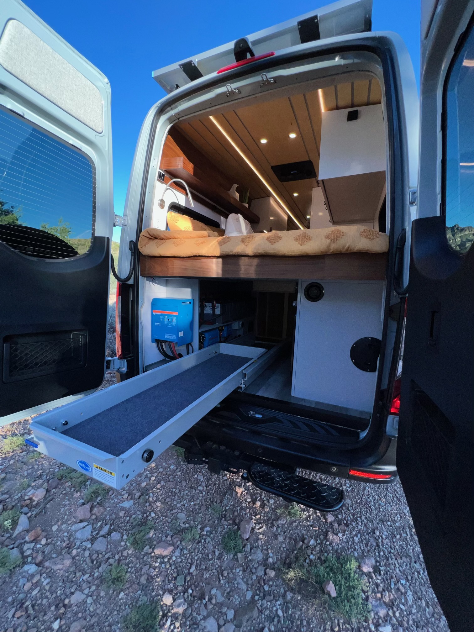 Inside an RV rental, open back doors reveal a camper van's cozy bed, pull-out storage, and Victron power setup.. Mercedes Sprinter Van 170 4x4 2022