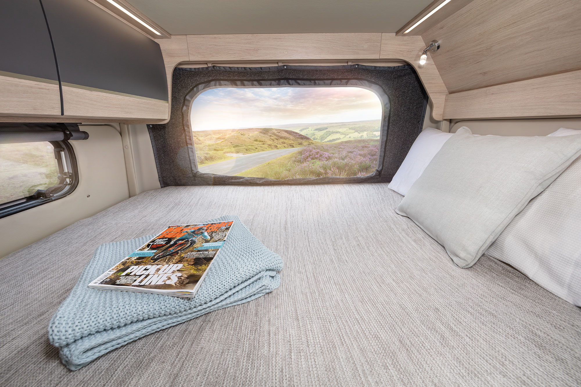 Comfortable RV rental interior with a neatly made bed, magazine, and an expansive mountain landscape view from the rear window.. Auto Trail Expedition 68 2025