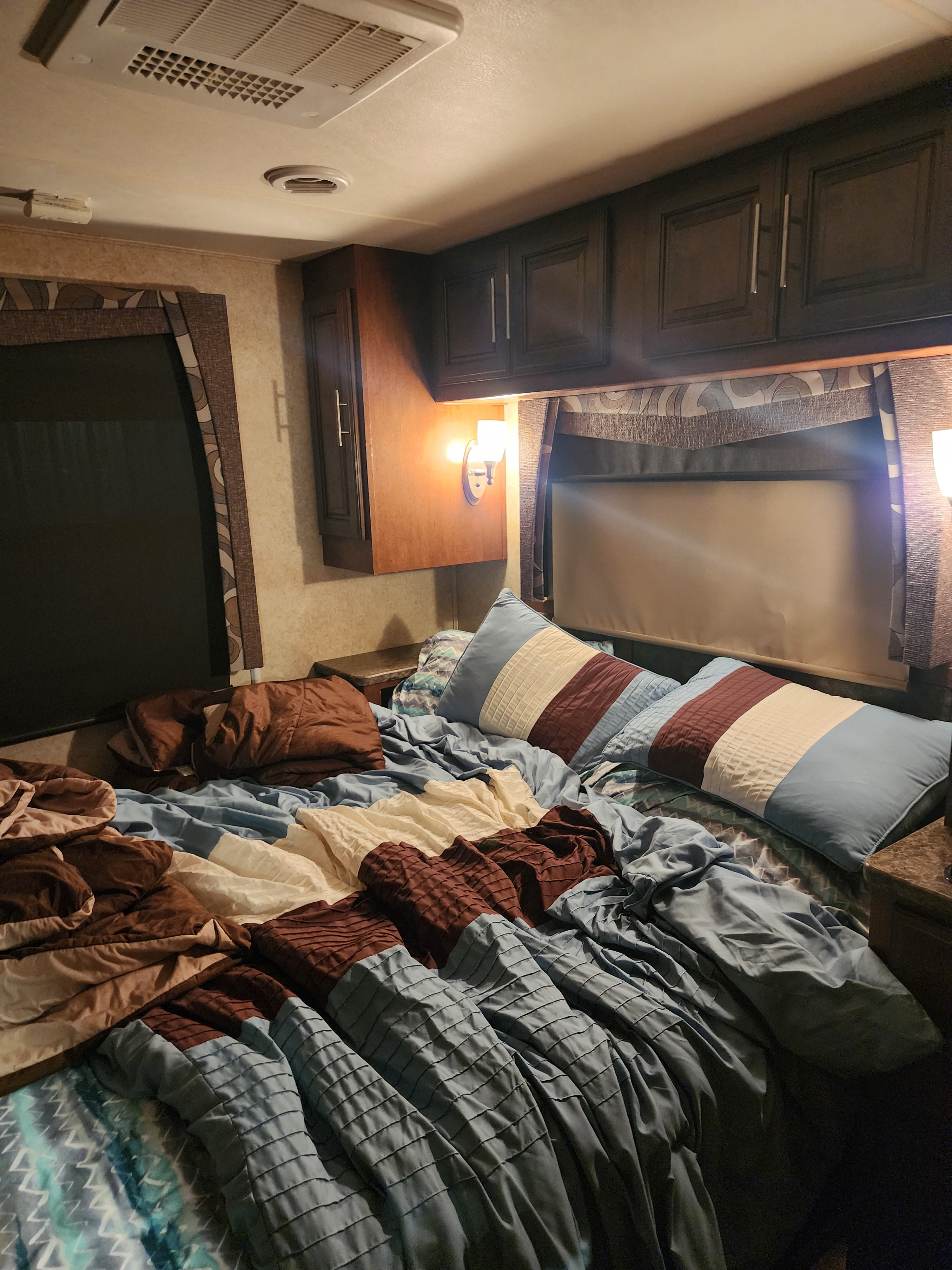 Cozy RV bedroom with unmade bed, blue/brown bedding, overhead cabinets, and bright sconce lights for an rv rental.. Forest River Georgetown 2015