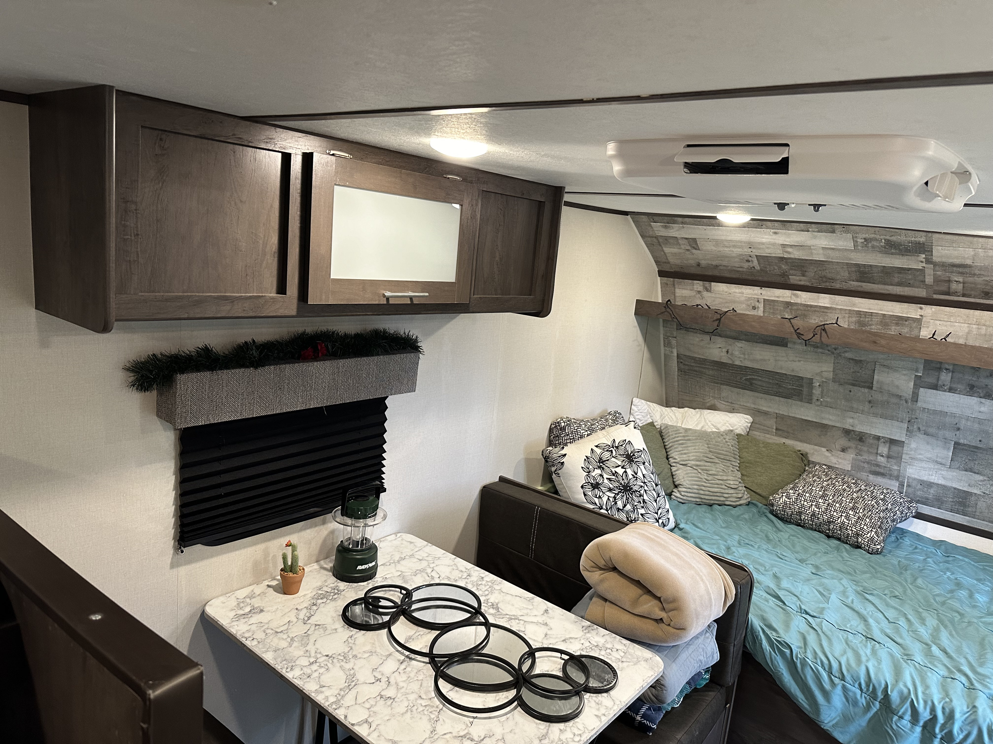 RV rental interior: cozy bedroom area with blue comforter, dining table, and dark wood overhead storage.. Coleman 17b 2022