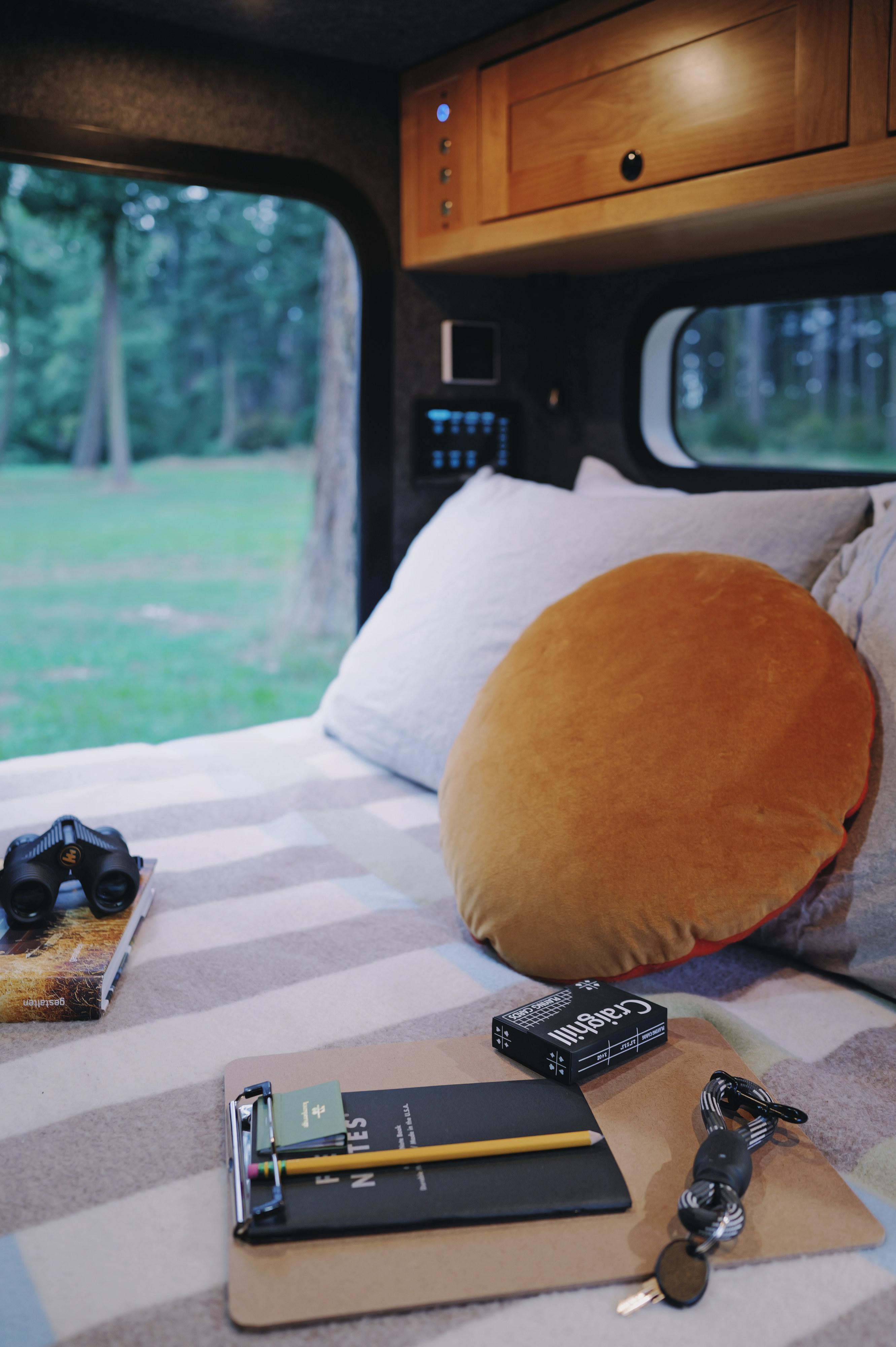 Cozy RV rental interior with a comfy bed, window view of nature, binoculars, a notebook, playing cards, and keys.. Campworks NS-1 Electrified 2024