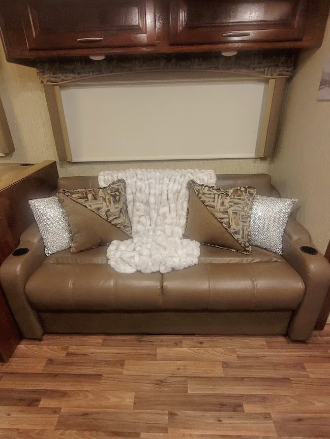Modern RV rental interior: brown sofa with patterned/sparkly pillows & fluffy white throw. Wood cabinets & window above.. Coachman Mirada 2018