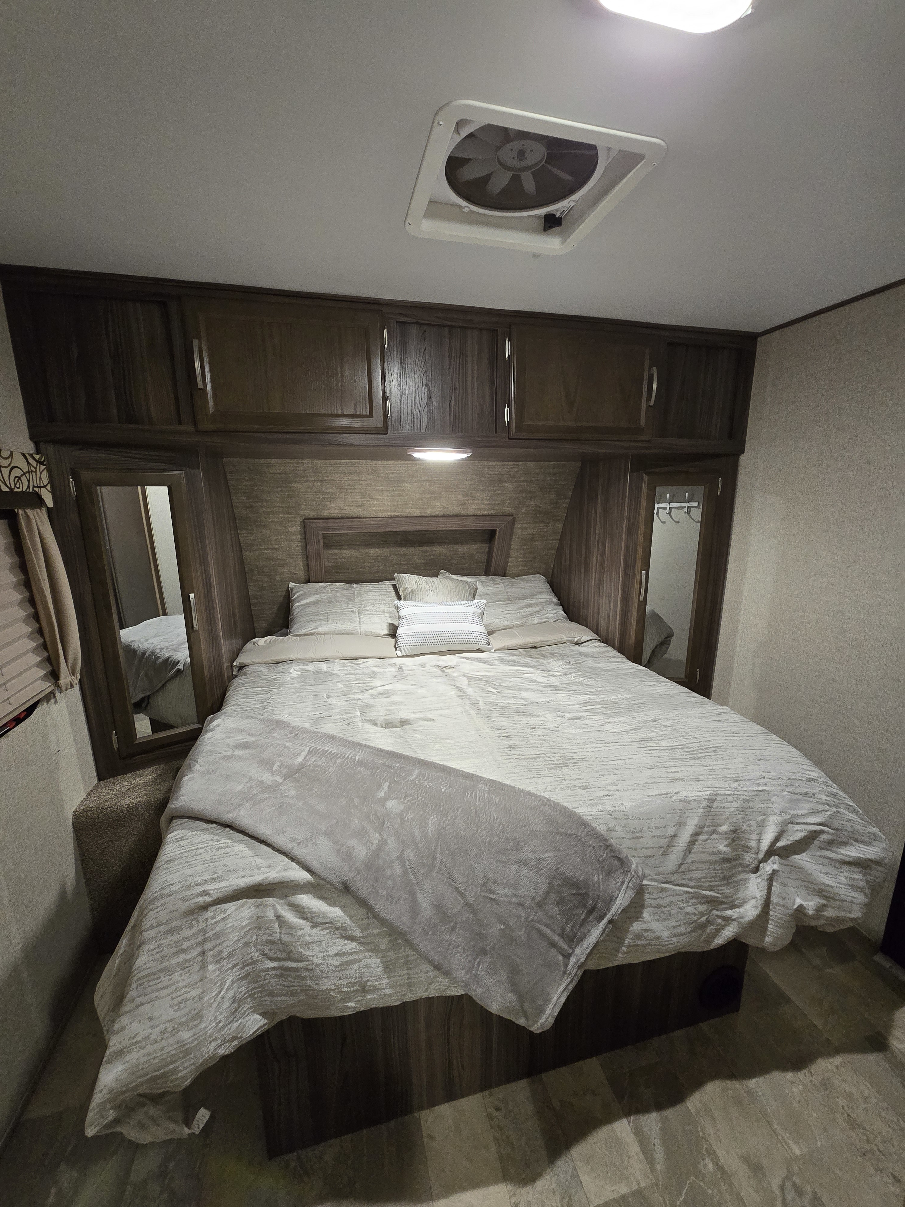 Spacious RV rental bedroom featuring a queen bed with neutral bedding, dark wood cabinets, a ceiling fan, and textured walls.. Coachmen Apex 2017