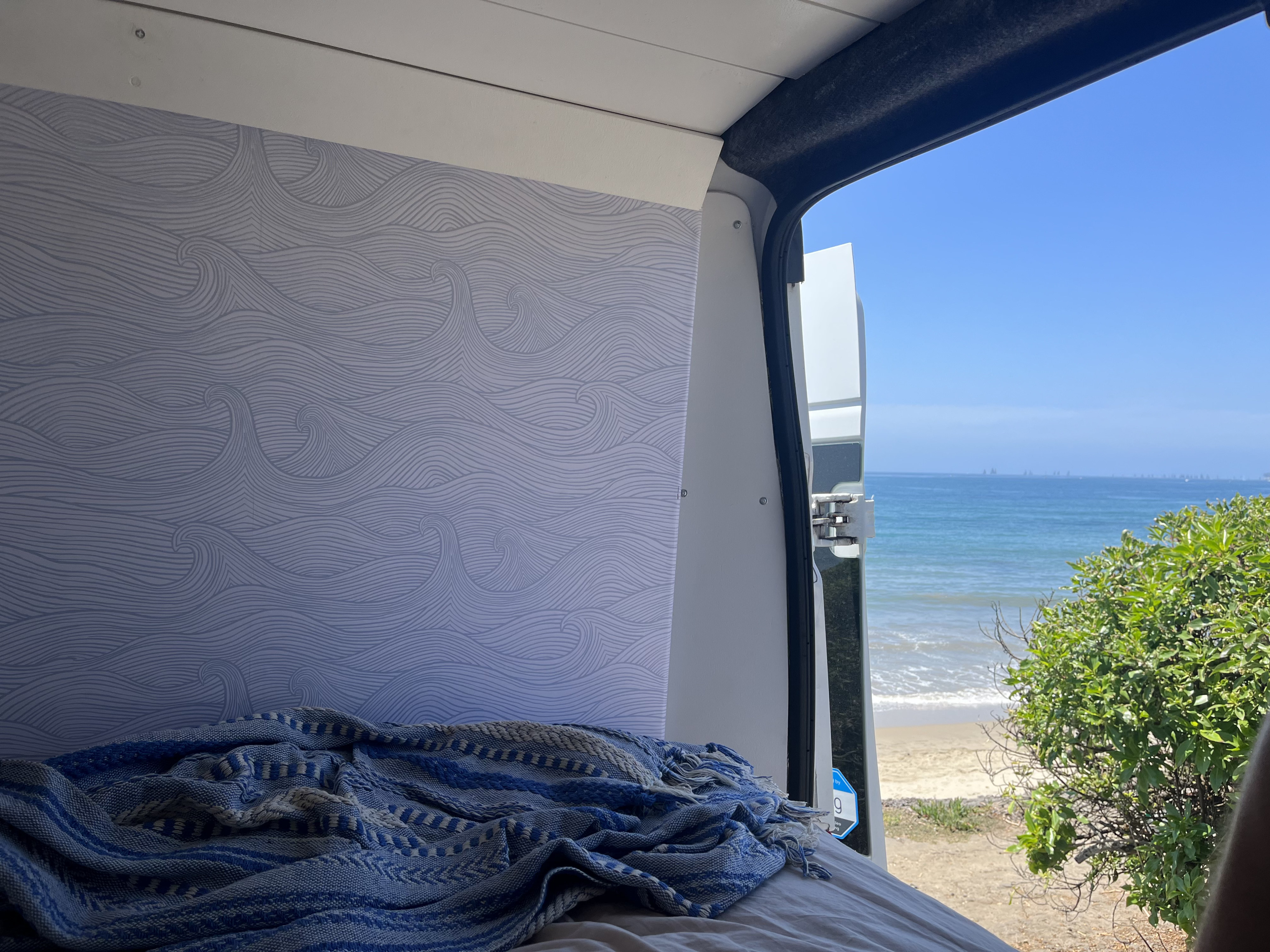 Enjoy the ocean view from this cozy RV rental's bed, featuring a wave-patterned wall. Ideal for beach travel and van life.. RAM Promaster 3500 2014