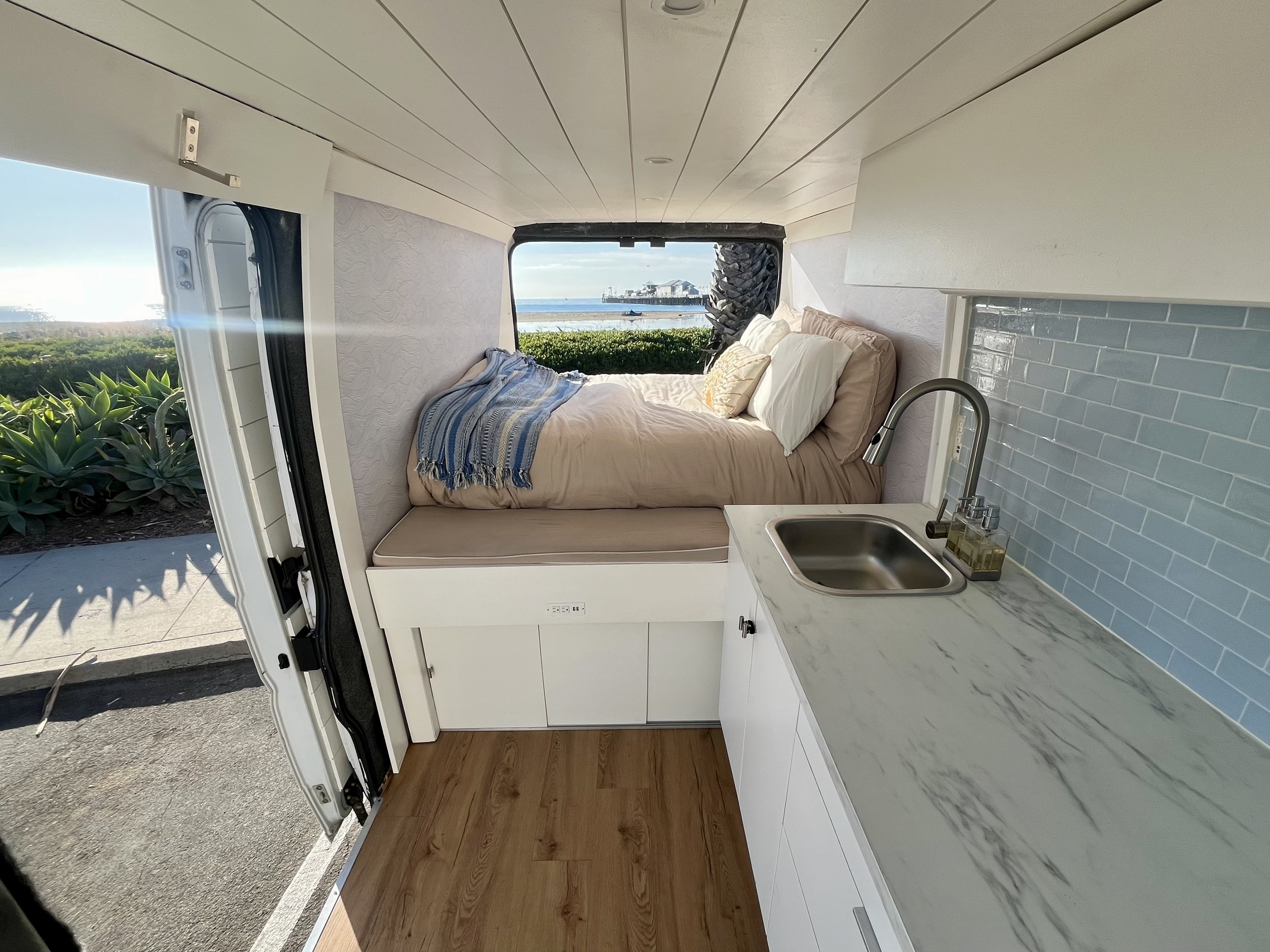 Stylish RV rental interior shows a cozy bed with ocean view, modern kitchen, and wood flooring in a bright camper van.. RAM Promaster 3500 2014