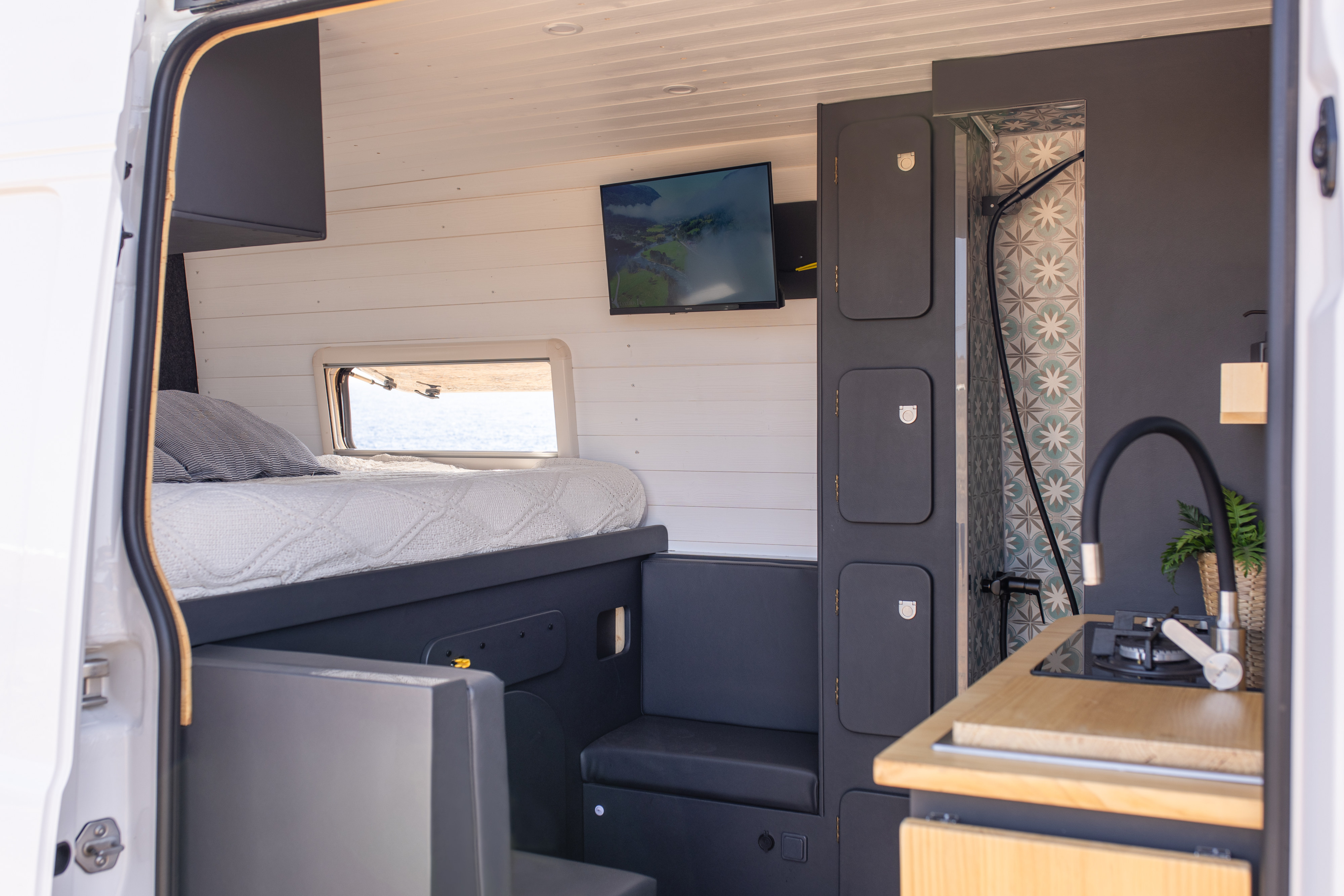 This modern RV rental interior features a cozy bed, TV, compact kitchen, and a patterned shower area, ideal for van life.. Volkswagen Crafter 2025