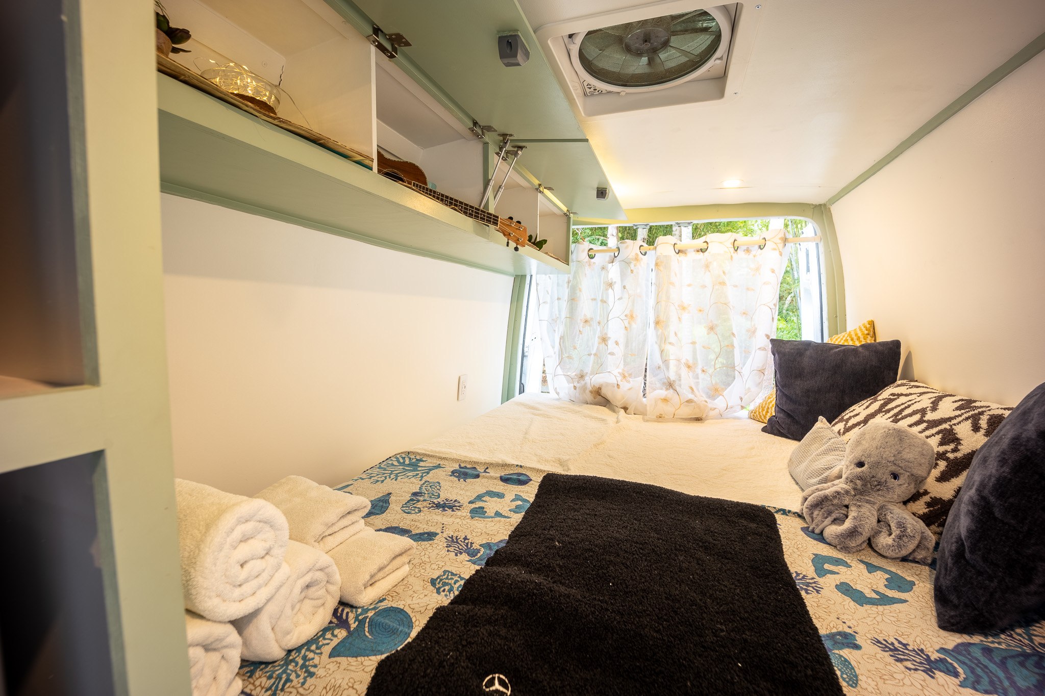 Cozy RV rental interior: bed, overhead storage with a ukulele, window with curtains, and ceiling fan.. Mercedes-Benz Sprinter 419 LWB High Roof 2013