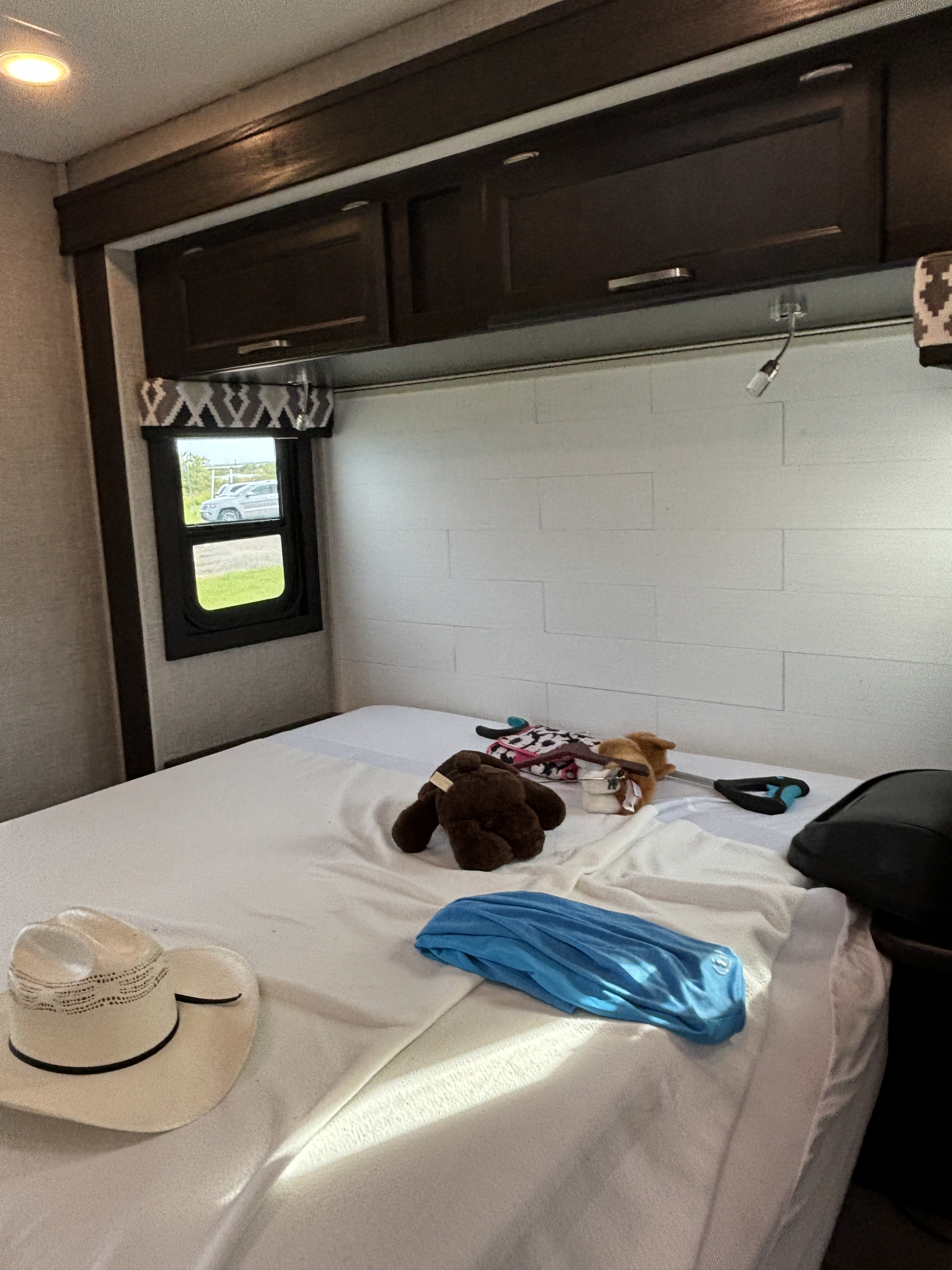 This RV rental bedroom features a bed with white sheets, a cowboy hat, toys, and a blue shirt; a window shows the outside.. Jayco Greyhawk 2022