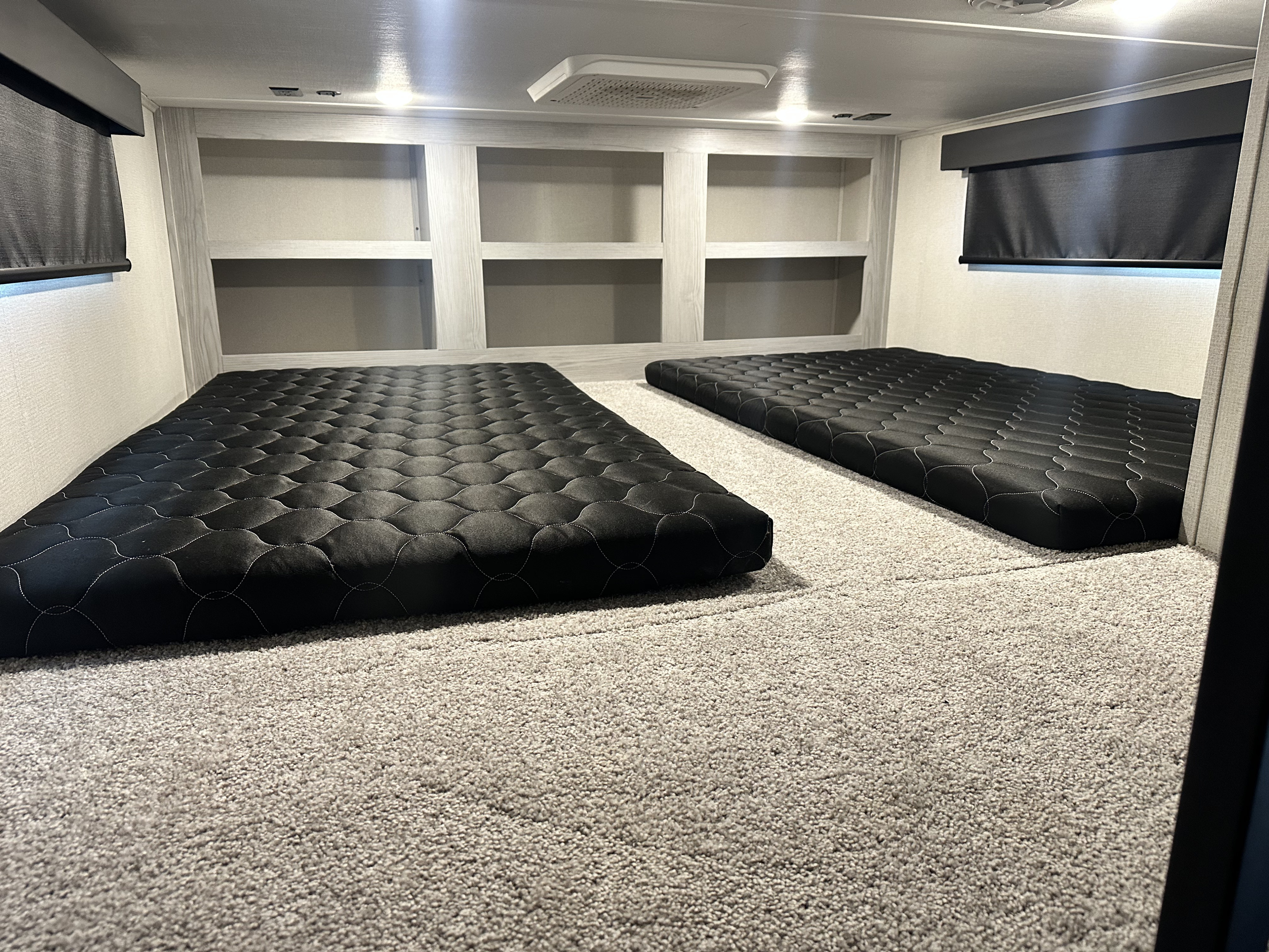 Inside an RV rental: a cozy sleeping area featuring two black quilted mattresses on carpet, with built-in shelves & window blinds.. Keystone RV Montana High Country 2023