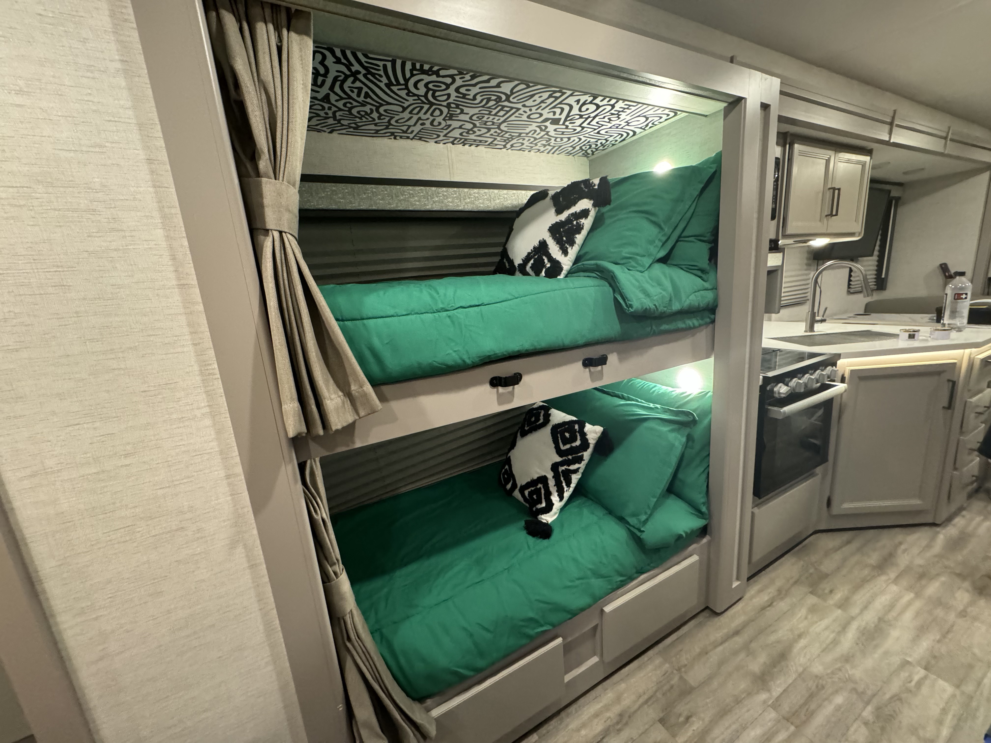 RV rental interior features two cozy bunk beds with bright green bedding, patterned ceilings, and a compact kitchen space.. Thor Resonate 2024