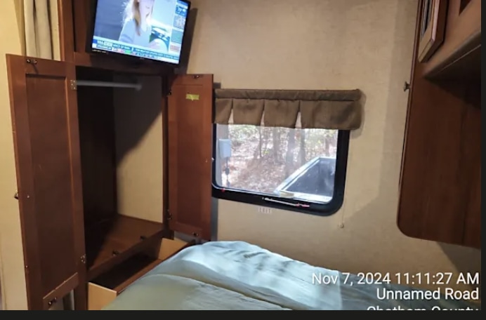 Inside an RV rental: a compact bedroom features a bed, mounted TV, open wardrobe, and window overlooking trees.. Heartland Mallard 2017