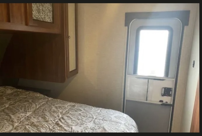 RV rental interior: patterned bed, wooden cabinets, and an entry door with a bright window & TV mount.. Heartland Mallard 2017
