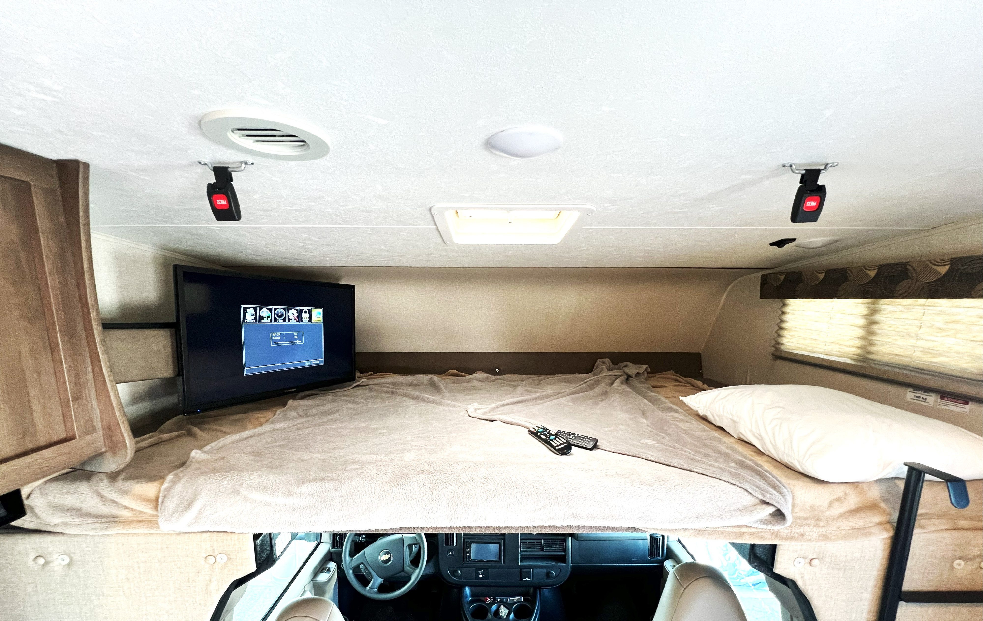 Inside an RV rental: a comfortable overhead bunk bed, TV, and remotes, with the cockpit visible below.. Coachman Freelander QB 2019